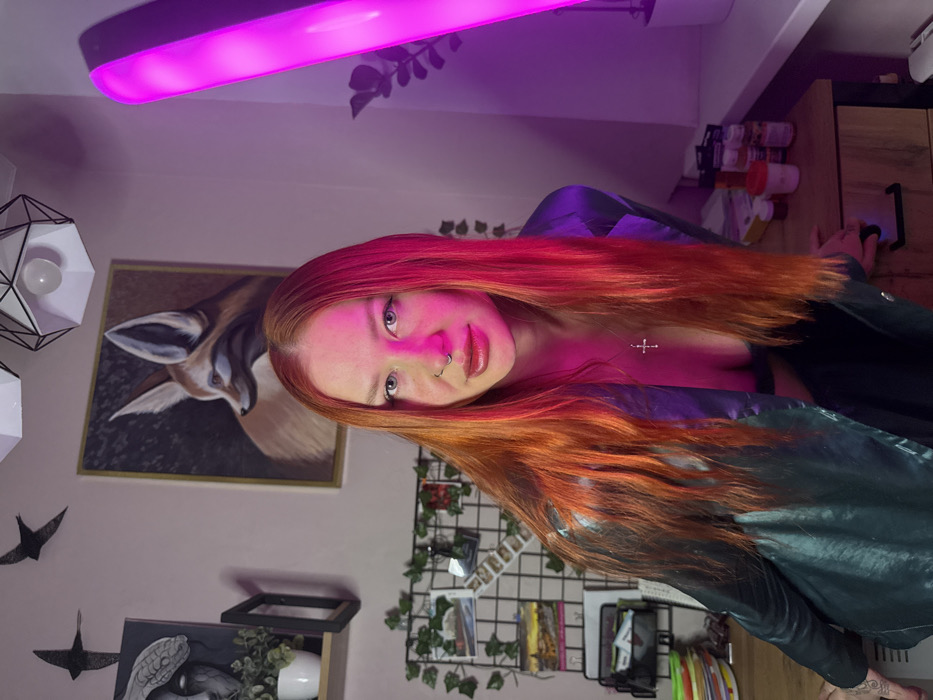 A woman with red hair, a septum piercing, and wearing a leather jacket poses for a photo in a room with pink lighting. Behind her is a painting of a deer, a metal grid with ivy, and a desk with supplies. Above is a pink light bar and a modern-style geometric light fixture. The woman is looking directly at the camera.