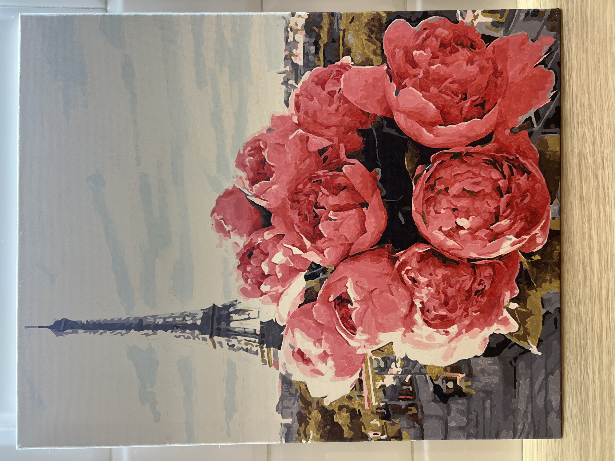 The image is a painting of a bouquet of pink flowers with the Eiffel Tower in the background. The sky is light blue, and the buildings below the flowers are various shades of gray and brown. There are no people depicted. The scene appears to be set in Paris, France, not Vnukovo, Russia. The painting seems to be a representation of Paris with a focus on the floral arrangement.