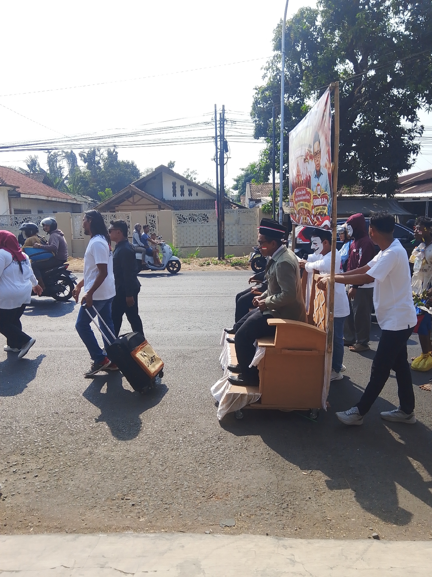 In Kalinyamatan, Indonesia, a group of people are participating in what appears to be a parade or procession. A large banner featuring a portrait of a man in glasses is displayed on a wooden structure, possibly a float. Several individuals are walking alongside or pushing this structure, with some wearing face paint or masks. One person is seated on the float and another is pulling a suitcase. In the background, houses and trees line the street, and other people on motorcycles are also visible. The scene suggests a public event taking place on a sunny day.