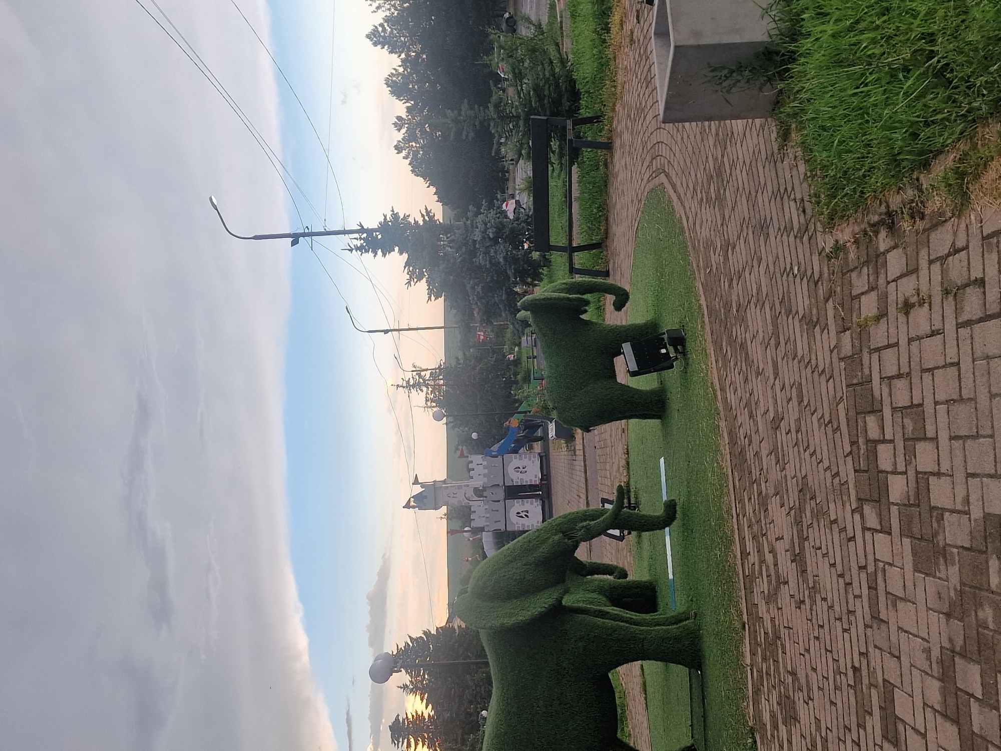 This image depicts an outdoor scene in Kursavka, Russia, featuring topiary elephant sculptures on a manicured lawn. In the background, a playground structure resembling a castle is visible, along with trees and a lamppost. The foreground is paved with brickwork, and a concrete planter is situated to the right. The sky is partly cloudy with a hint of sunset. No people are clearly visible in the image.