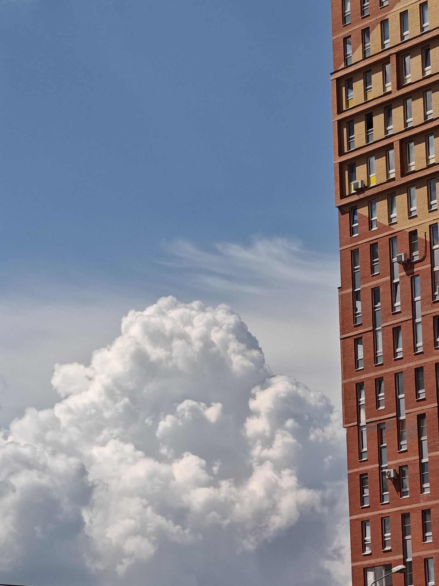 In the city of Shcherbinka, Russia, a close-up, low-angle shot captures a modern brick apartment building partially obscuring a vast blue sky filled with dramatic, voluminous white clouds. The building, constructed with reddish-brown bricks and accented with lighter-colored bricks and yellow window frames, dominates the right side of the frame. Its facade features numerous rectangular windows, many with reflective glass, arranged in a grid pattern. Several air conditioning units are visible mounted on the exterior walls. To the left of the building, a massive, cumulonimbus cloud formation with a dense, puffy texture and shadowed undersides occupies the majority of the sky. Wispy, lighter clouds are present higher in the sky, adding depth and texture to the celestial backdrop. The lighting suggests a sunny day, with strong shadows cast by the building.