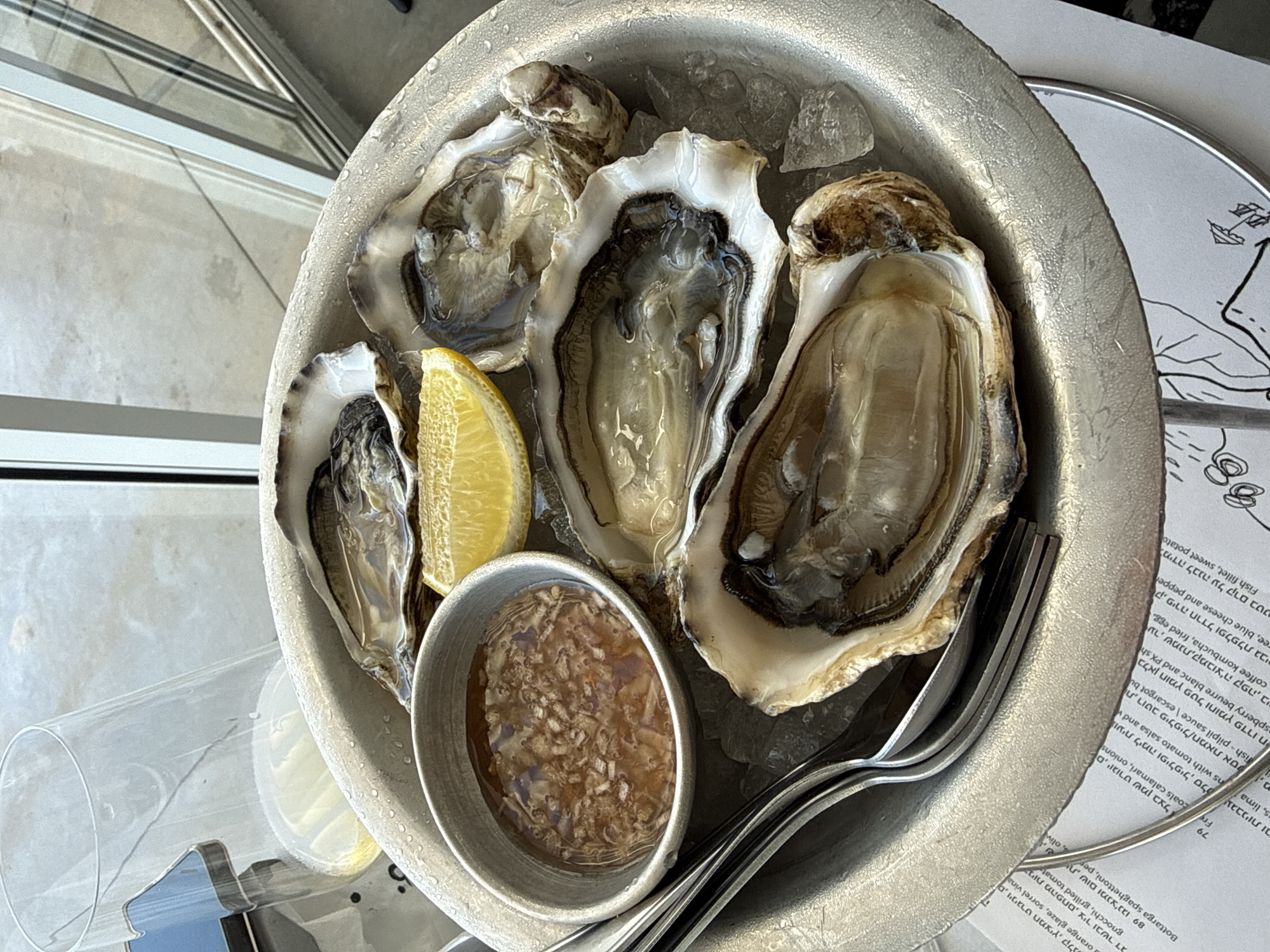The image presents a close-up, high-angle view of a metallic serving bowl filled with oysters on ice, a lemon wedge, and a small container of sauce. Utensils are partially visible within the bowl as well. The oysters are in their shells, with visible meat and liquid. A portion of a menu with Hebrew text is visible in the background, as well as restaurant glassware. Considering the Hebrew text on the menu, it's plausible that the photo was taken in Tel-Aviv, Israel.
