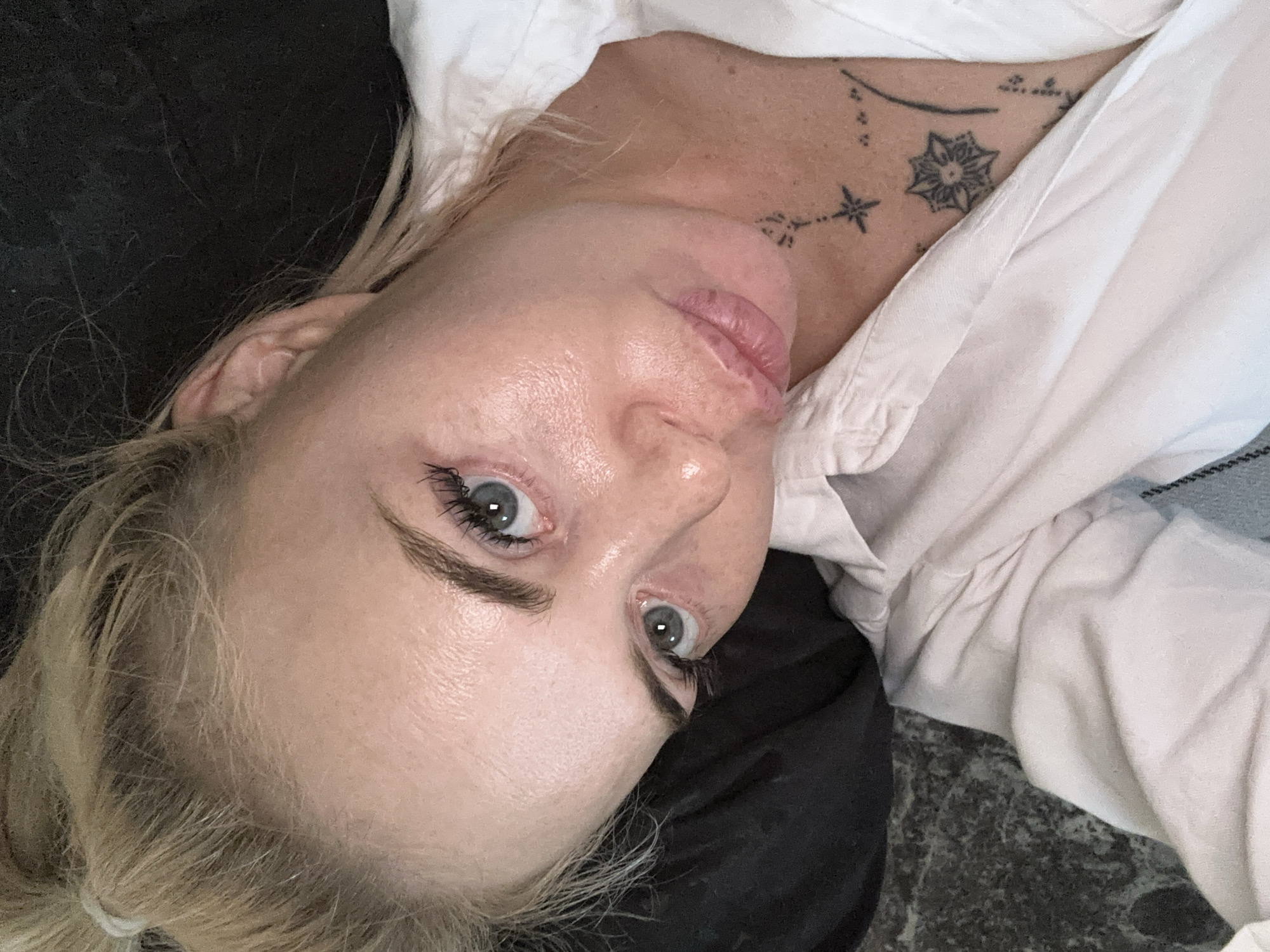 A blonde woman with blue eyes and light skin is lying down, looking up at the camera. She is wearing a white collared shirt that is partially unbuttoned, revealing tattoos on her chest. Her hair is pulled back in a messy bun. The background is dark and out of focus. The overall lighting is soft and diffused.