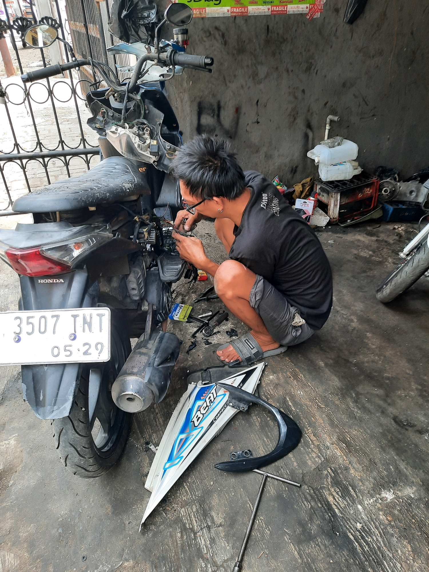 In East Jakarta, Indonesia, a person is seen squatting down to repair a motorcycle. The mechanic, wearing a black t-shirt and grey shorts, has glasses on and is focused on the wiring of the motorcycle. The motorcycle is a dark blue scooter with a license plate that reads "3507 TNT 05.29". Various tools and motorcycle parts are scattered on the grimy concrete floor, indicating an outdoor repair shop or a workshop. In the background, a concrete wall is visible, along with some stacked containers and debris. The overall scene depicts a typical urban setting for motorcycle maintenance and repair.