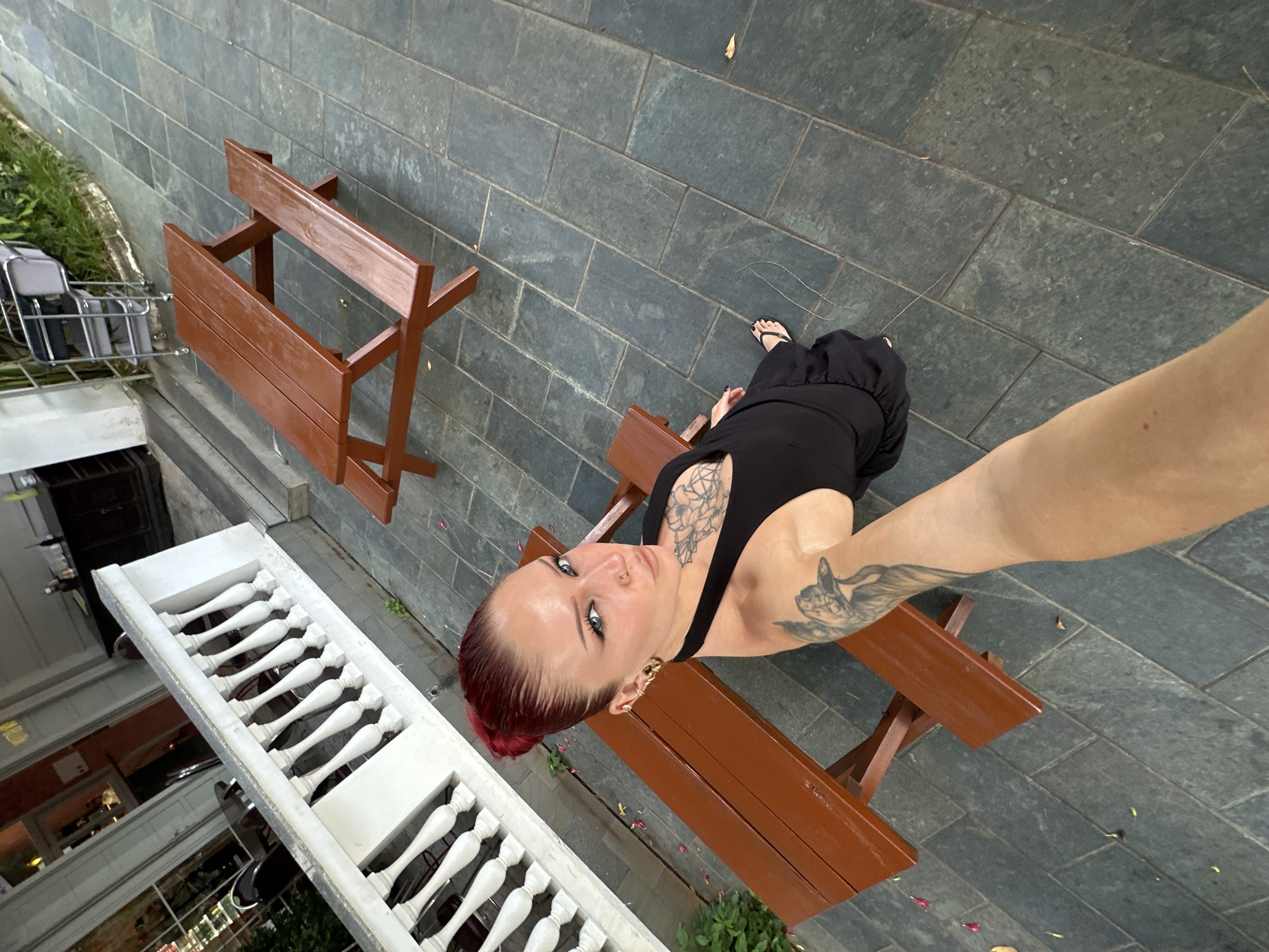 A young woman with bright red hair pulled into a bun, wearing a black halter dress and flip-flops, poses for a selfie outdoors in Hoi An, Vietnam. She has extensive tattoos on her arms and chest, and is wearing large hoop earrings and a nose ring. She is seated on a wooden bench next to a wooden picnic table. The setting appears to be a courtyard or patio with dark, grey stone tiles on the ground. In the background, there is a white railing with decorative spindles, and beyond that, a building with large windows and doors, suggesting a restaurant or cafe. There is also another picnic table visible further in the background. The lighting suggests it is daytime, possibly late afternoon or early evening.