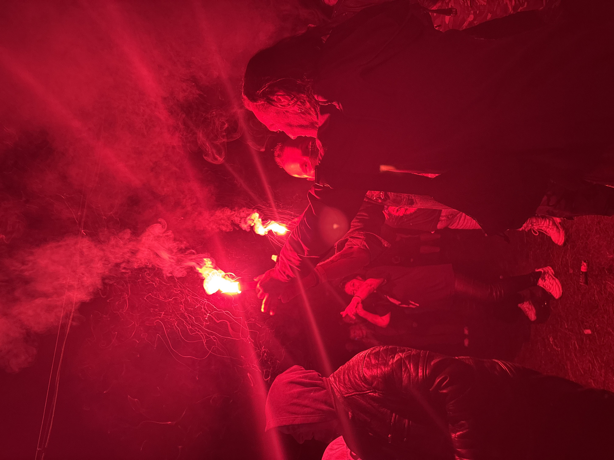The image depicts a nighttime outdoor gathering in Большая Кивара, Russia, characterized by intense red illumination and dense smoke. Multiple individuals are present, obscured and highly saturated by the red light.
In the foreground, to the left, a person with their back largely to the viewer wears a dark, possibly leather, hooded jacket with textured detailing. Their head is turned slightly to the right, observing the scene. To their right, in the mid-ground, another person appears to be holding a white or light-colored object, possibly a megaphone, up to their mouth.
The central action involves several individuals holding and activating pyrotechnic devices, likely flares or smoke bombs. One prominent figure, positioned right of center, holds a device upwards with their right hand, emitting bright yellow-orange flames and a substantial volume of red smoke and sparks. This individual is seen in profile, with long hair visible. Another hand, either belonging to this person or someone in close proximity, is also seen holding an additional device. Further to the right, a third individual is partially visible, also contributing to the light and smoke with another flare.
The entire scene is enveloped in a thick, red smoke or haze, which is particularly dense in the upper and mid-sections of the frame, obscuring the background. Fine, glowing red streaks and embers are visible throughout the smoky atmosphere. The ground, visible in the lower portion of the image, appears dark and uneven, with scattered discarded items such as an empty can or bottle evident in the bottom right corner. The clothing of the participants appears casual, including sneakers visible on some individuals in the lower right. The overall impression is that of an active, crowded event under conditions of low natural light and strong artificial red light.