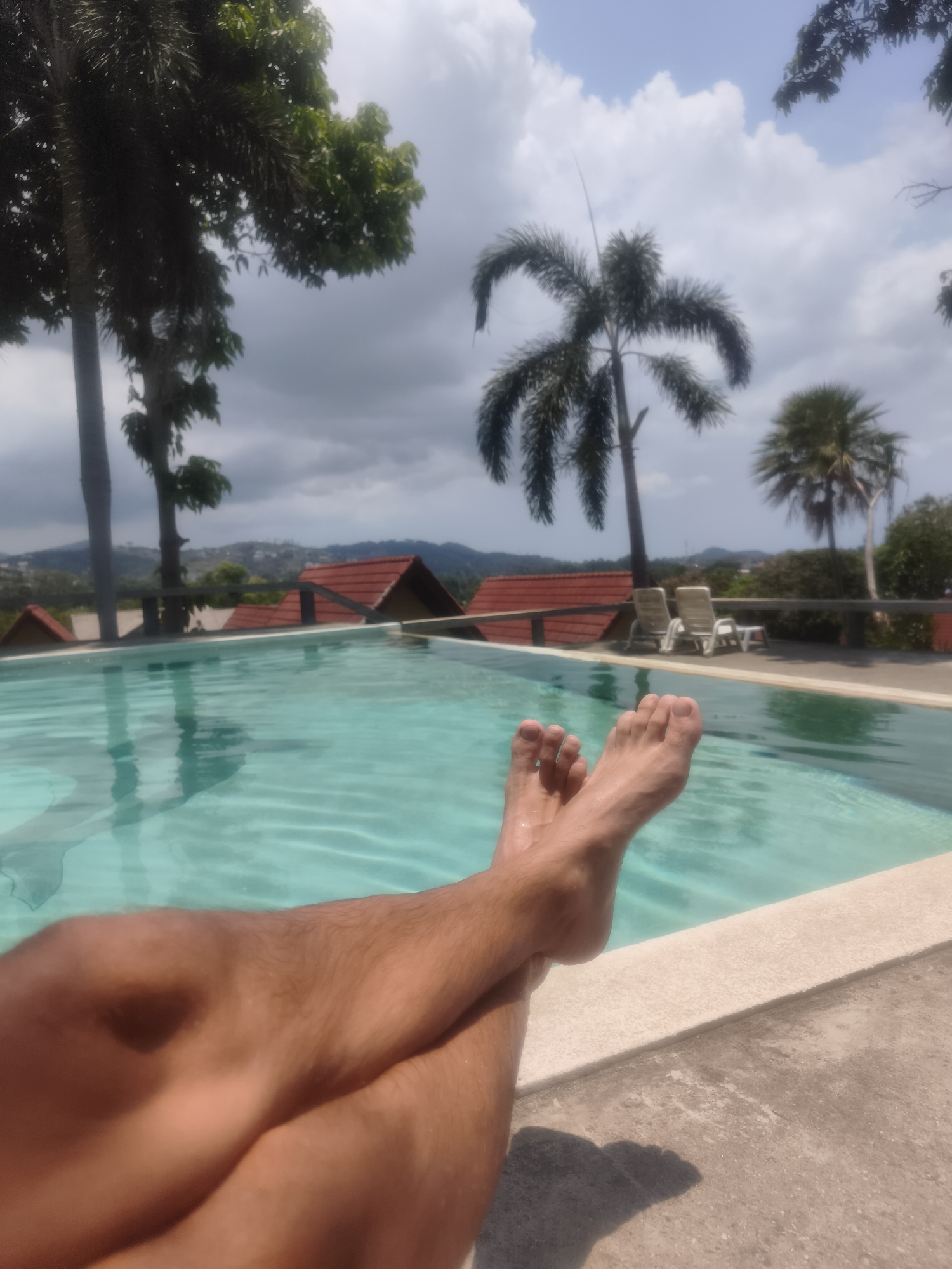 The image captures a first-person perspective of someone relaxing poolside on a sunny day in what appears to be a tropical location. The main subject is a person's legs and bare feet, crossed at the ankles and extended towards a shimmering turquoise swimming pool. The legs and feet show some tan lines and body hair. The person is likely sitting or lounging at the edge of the pool, with the water's surface reflecting sunlight and showing gentle ripples.
Beyond the pool, the scene unfolds with a picturesque view. Lush green palm trees, along with other tropical foliage, frame the upper and left portions of the image against a bright sky. The sky is partly cloudy, with scattered white clouds contrasting against a lighter blue. In the middle ground, several houses or bungalows with distinctive red-tiled roofs are visible, nestled amidst the greenery. Further in the distance, hills or mountains create a natural backdrop. Two white lounge chairs are unoccupied on the poolside patio, suggesting a resort or vacation setting.
The overall impression is one of leisure and tranquility. The time of day is clearly daytime, characterized by bright illumination, although the presence of clouds suggests a mix of sun and shade. There is no visible text in the image. The setting, characterized by palm trees, red-roofed buildings, and distant hills, aligns with the provided location context of บ้านบางรักษ์, Thailand, suggesting a scenic, possibly mountainous or hillside resort area.