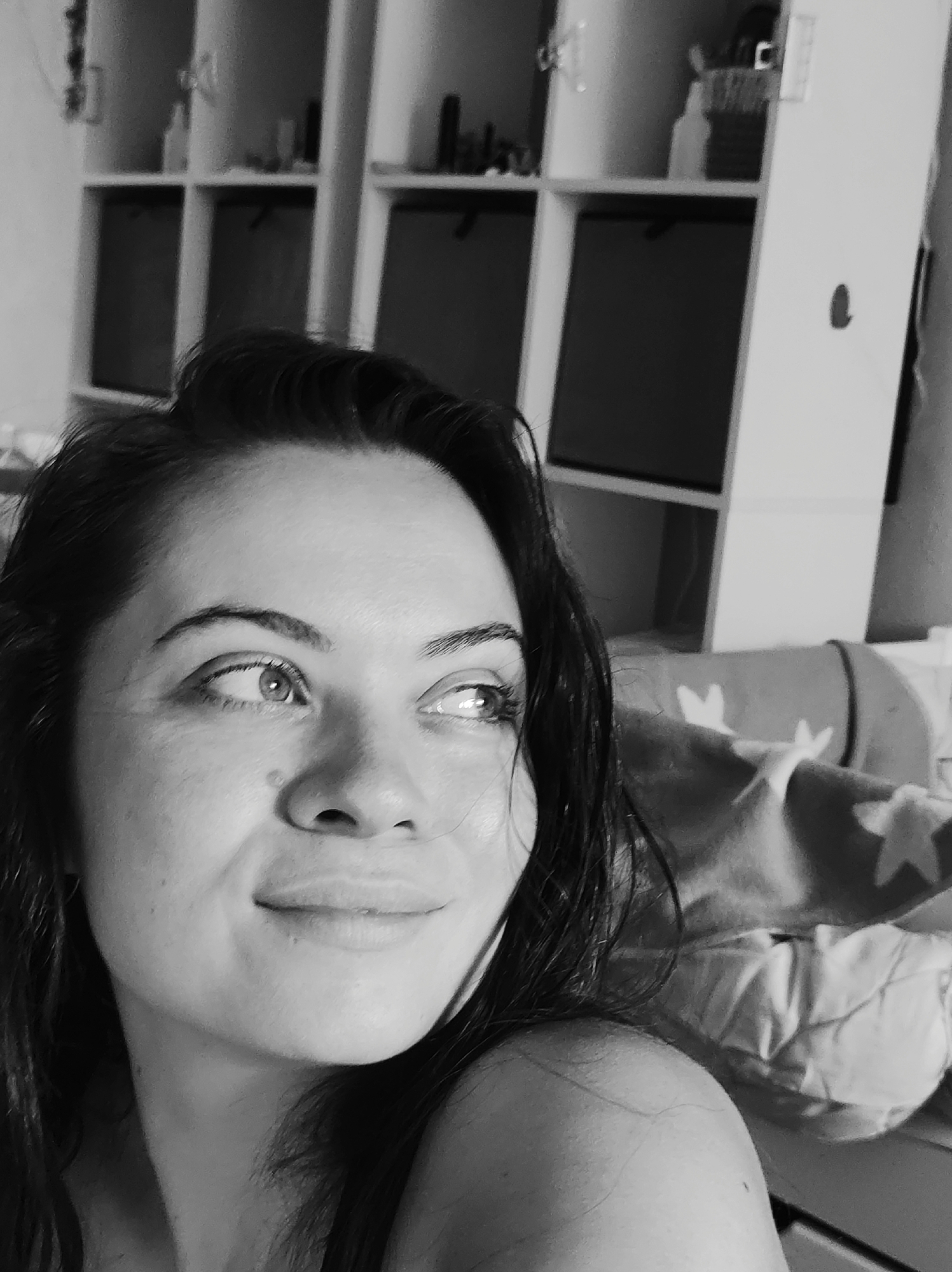 The black and white, close-up, waist-up photograph shows a young woman looking up and to the right. She has dark hair and fair skin. She is wearing a dark colored top. The background consists of a white shelving unit with various items on the shelves. To the right of the woman, a blanket with star patterns is visible. The image appears to be taken indoors. The scene does not provide any specific visual cues that would allow for the identification of the location within the city of Chaikovsky, Russia.