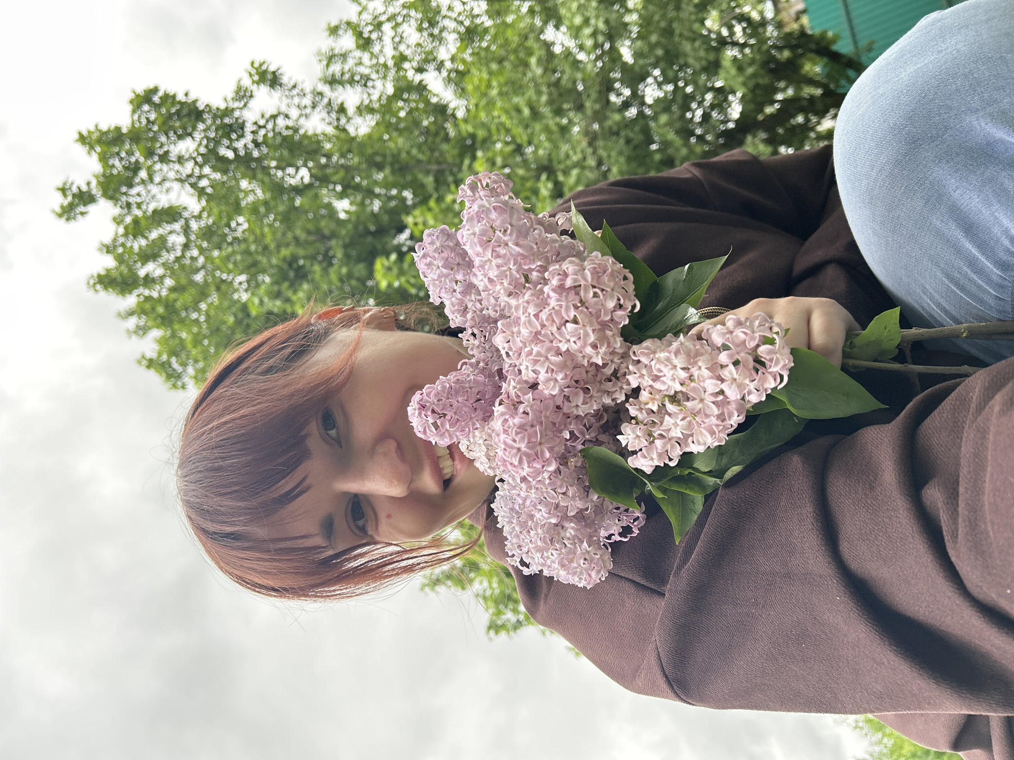 A woman with auburn hair and bangs smiles at the camera, holding a bouquet of lilac flowers. She wears a brown hoodie and blue jeans. The background consists of green trees and a cloudy sky. The location appears to be Фоки, Russia.
