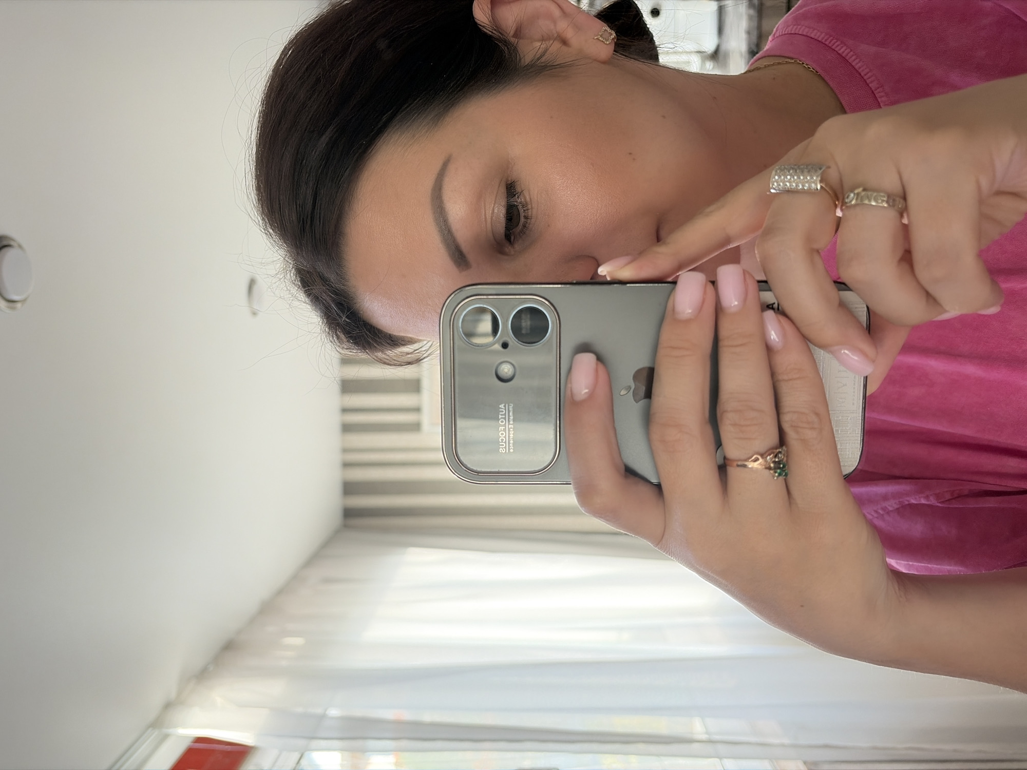 A woman, with dark hair pulled back and a pink shirt, holds a gray smartphone to take a selfie. She is wearing two rings, one with a green stone and the other with a diamond band. A small, gold earring shaped like a cat is visible on her right ear. The background shows a window with white curtains and a wall with vertical stripes. The scene appears to be indoors, possibly in an apartment or hotel room in Sochi, Russia.