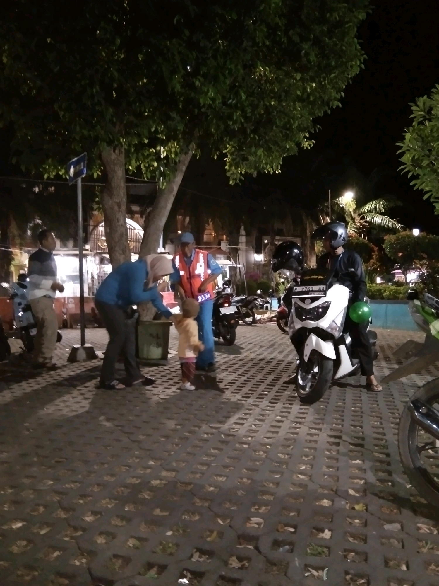 In Bondowoso, Indonesia, at night, a group of people are gathered near trees and motorcycles. A man in a reflective vest is interacting with a child, who is wearing a peach-colored shirt and striped pants, reaching out a hand towards the man. A woman in a blue jacket and headscarf is bent over, possibly assisting the child. To the left, a man in a striped sweater and light-colored pants stands near a scooter. In the foreground, a white motorcycle with a license plate reading "P 2763 AA" is parked, with a person wearing a helmet seated on it, holding a green balloon. Other motorcycles are visible in the background. The ground is paved with interlocking concrete blocks, with some debris and fallen leaves scattered on it. The scene appears to be in a public outdoor space, possibly a park or roadside area, illuminated by artificial lighting and streetlights.