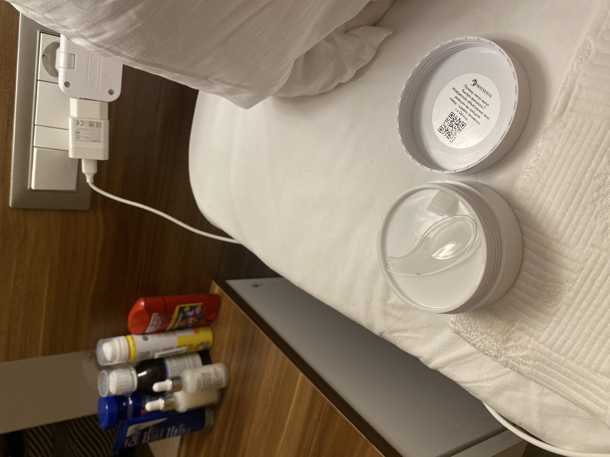 The image depicts a close-up, indoor scene likely in a hotel room in Aksu, Turkey. The foreground is dominated by a white bed with a white duvet, on which rests an open white jar containing eye patches. A clear plastic spatula is placed on top of the eye patches inside the jar. Next to the jar, on the bed, is the white lid of the jar, which displays a QR code and text in Russian, including a brand name "MATSESTA".
To the left, a wooden nightstand is visible, holding several toiletries. These include a blue tube, a dark-colored bottle with a white cap, two small bottles with droppers and white caps, a yellow bottle with a white cap, and a red bottle with a white cap.
In the background, a white electrical outlet with multiple switches and a white phone charger plugged into it is mounted on a wooden wall. The charger has text and symbols, including "MADE IN CH". A white cable extends from the charger, partially visible. The overall impression is a neatly arranged bedside setup, with personal care items and charging electronics. The lighting appears to be artificial and warm.