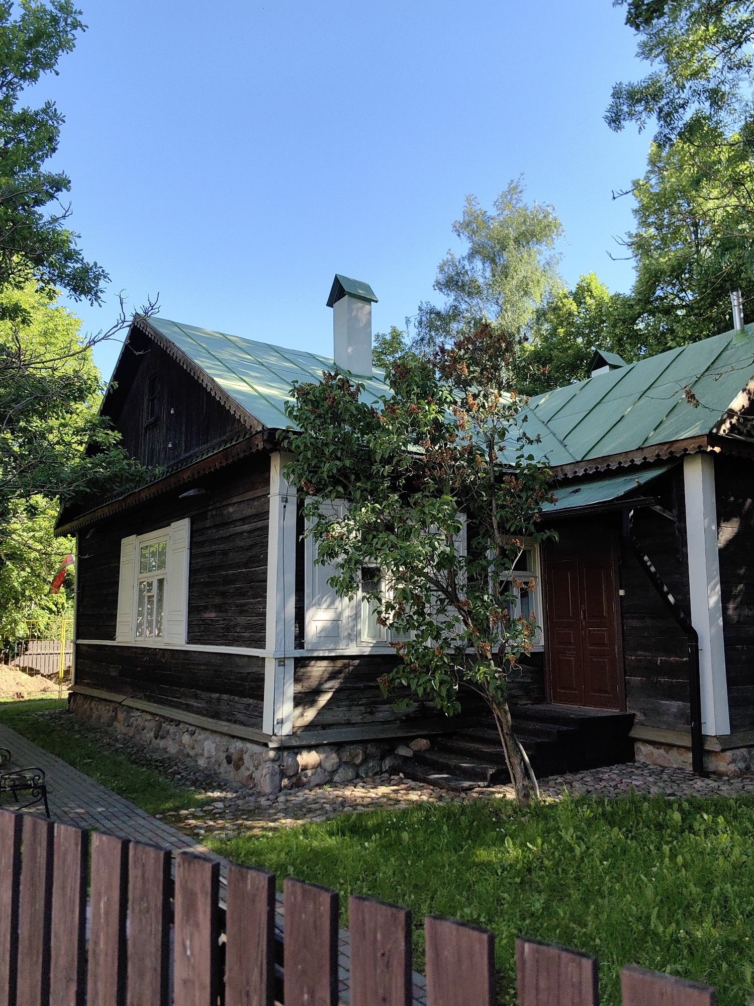 A dark wooden house with a green metal roof is shown from a low angle, with a brown wooden fence in the foreground. The house has white-framed windows with shutters, a stone foundation, and a chimney. A small tree with sparse leaves stands in front of the house, and a brown wooden door is visible on the right side. The scene is set in a grassy area with trees surrounding the house, under a clear blue sky. This house is located in the ethnographic complex "Belarusian Fable" in Minsk, Belarus. There are no people visible in the image.