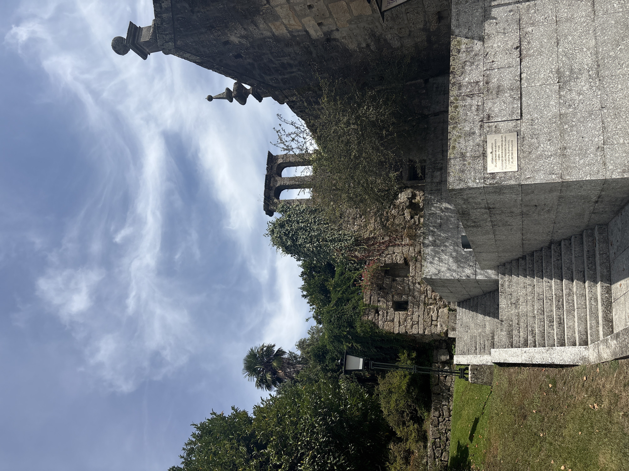 This image depicts the Largo do Toural in Guimarães, Portugal. A stone staircase with granite steps ascends from the lower left of the frame towards a partially ruined stone structure overgrown with ivy and foliage. To the right of the staircase, a large, textured granite wall features a commemorative plaque. In the background, more ancient stone ruins with arched openings are visible, also draped in greenery. Lush trees and a palm tree frame the scene on the left and behind the ruins. The sky above is a light blue with wispy white clouds. A street lamp stands beside the staircase. The overall impression is one of historical architecture blending with nature.
