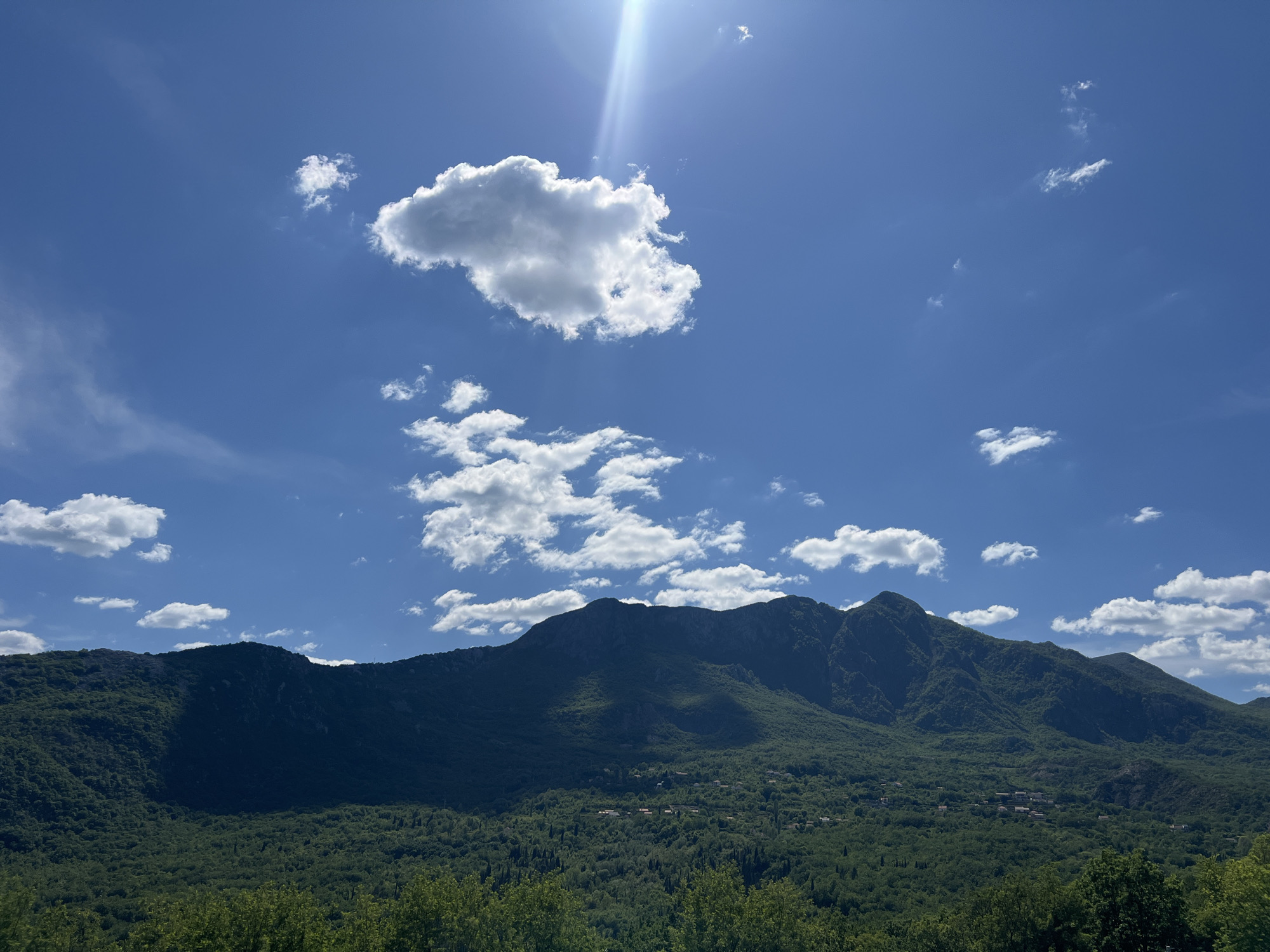 The image shows a daytime view of Sotonići, Montenegro. The foreground features a dense forest. A mountain range dominates the middle ground, its slopes covered in lush greenery. Scattered buildings are visible amongst the trees on the lower slopes. The sky is bright blue with scattered cumulus clouds. The sun is visible in the upper part of the image, causing lens flare.