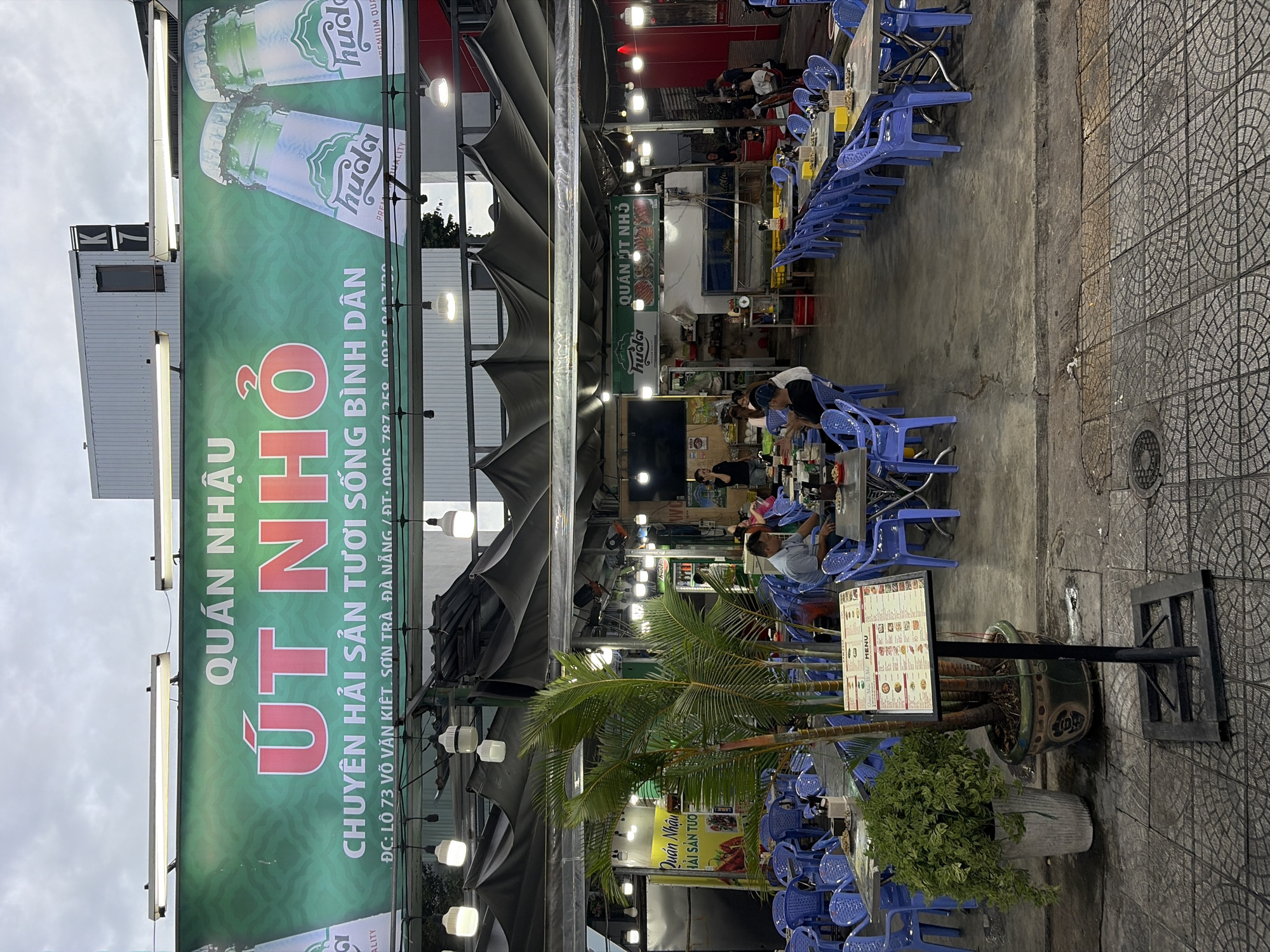 The image depicts the exterior of a busy outdoor restaurant in Da Nang, Vietnam, identified by the signage which reads "QUÁN NHẬU ÚT NHỎ" (Ut Nho Eatery) and the address "ĐC: LÔ 73 VÕ VĂN KIỆT, SƠN TRÀ, ĐÀ NẴNG". The restaurant has a large, green awning with the establishment's name prominently displayed, along with images of two beer bottles, identified as "Huda".
The seating area is set up with numerous blue plastic chairs arranged around metal tables. Many tables are occupied by patrons, suggesting a lively dining atmosphere. In the foreground, a large potted palm tree stands next to a menu stand displaying a Vietnamese menu. The ground is paved with a patterned concrete surface. The overall environment is characteristic of an urban dining setting in Vietnam, likely in the evening given the ambient lighting from overhead lamps.