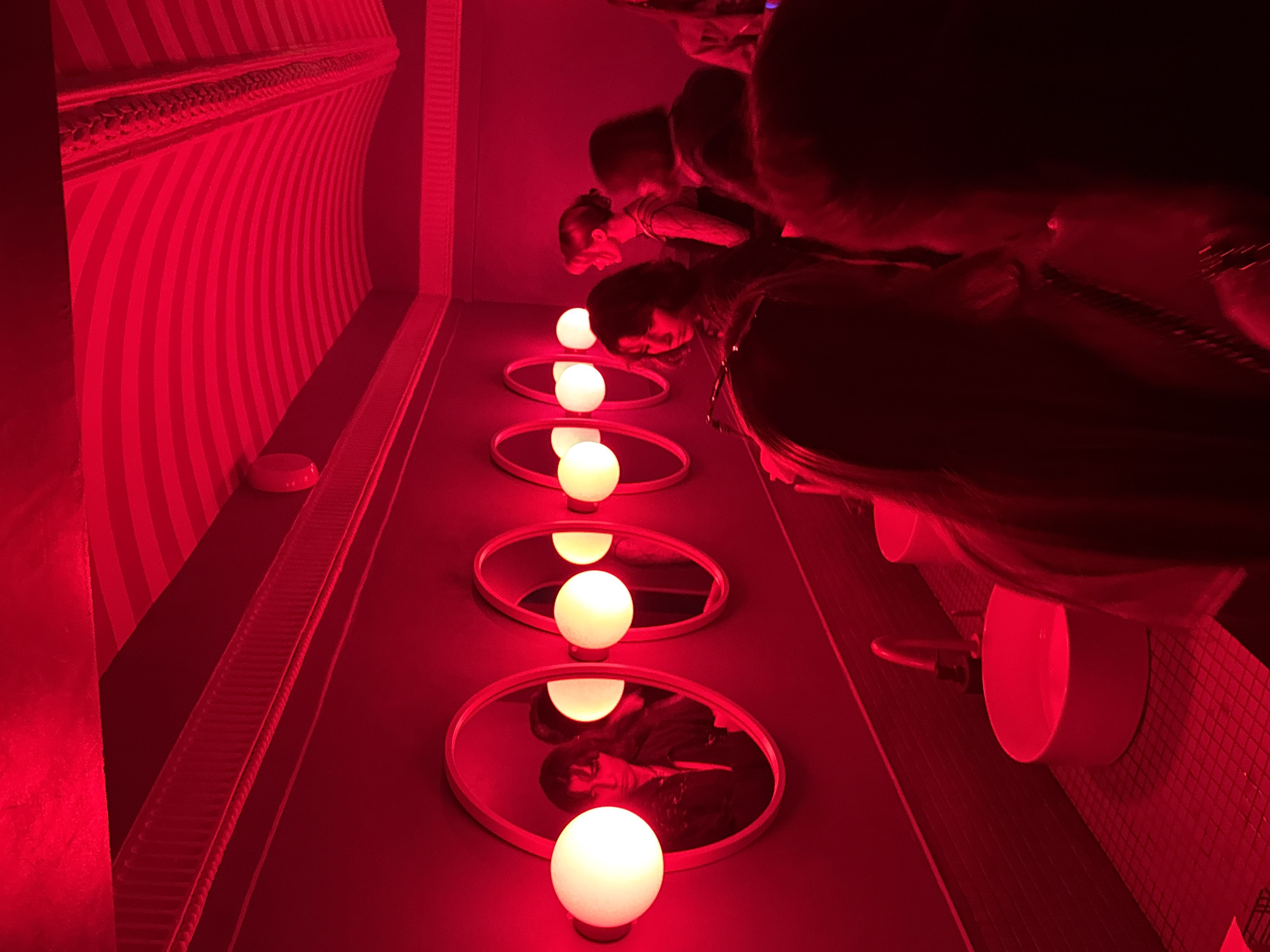The image depicts an interior scene of a public restroom, specified as located in Укромновское сельское поселение, Ukraine. The entire environment is saturated with a dominant red light, creating a dramatic and low-light atmosphere.
The left wall features a series of five oval mirrors, each flanked by a spherical, warm white light fixture. These lights provide the only distinct non-red illumination in the scene. Below the mirrors, a dark counter holds at least two modern, cylindrical white basin sinks, with a metallic faucet visible above the nearest one. The floor beneath the sinks is tiled with small, dark squares, which appear reddish due to the ambient lighting.
The ceiling on the right side of the room exhibits a distinct architectural feature: a vaulted or arched design with alternating red and white stripes. Decorative molding or trim with a ribbed pattern runs along the upper perimeter of the walls.
Multiple individuals, predominantly women, are present in the restroom. In the foreground, the back and shoulder of a person, wearing a dark garment with a visible dark strap or chain, partially obscure the right side of the frame. Reflected prominently in the first mirror on the left is a person with dark, shoulder-length hair, wearing a dark jacket, illuminated by the adjacent globe light. Further back in the mid-ground, a person with dark hair stands near the sinks, facing towards the mirrors. Behind them, another individual with light-colored hair, styled in an updo, is seen in profile, looking towards the right, wearing a patterned top. A fourth person, largely seen from behind, is situated near this individual. Additional blurry figures are discernible in the background, suggesting more people are present deeper within the room. Individuals appear to be either using the facilities or waiting, with no explicit interactions visible between them.