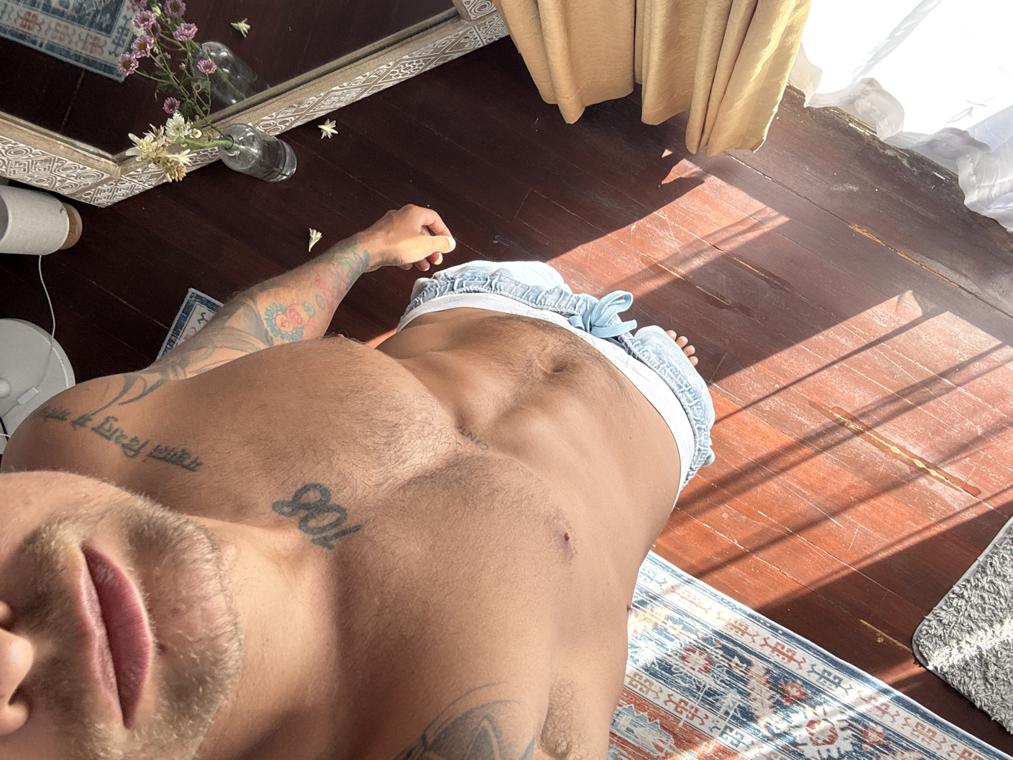 A close-up, overhead shot captures a man from the chest down, standing on a dark wooden floor. He is wearing light blue denim shorts with a white waistband visible. Sunlight streams in from the right, casting long shadows across the floor and highlighting a patterned rug on the left. A portion of a large mirror with a textured wooden frame is visible on the right, reflecting a small vase with flowers. The man has tattoos on his chest and arm, with the number "108" tattooed on his upper chest. He is shirtless, revealing his abdominal muscles.