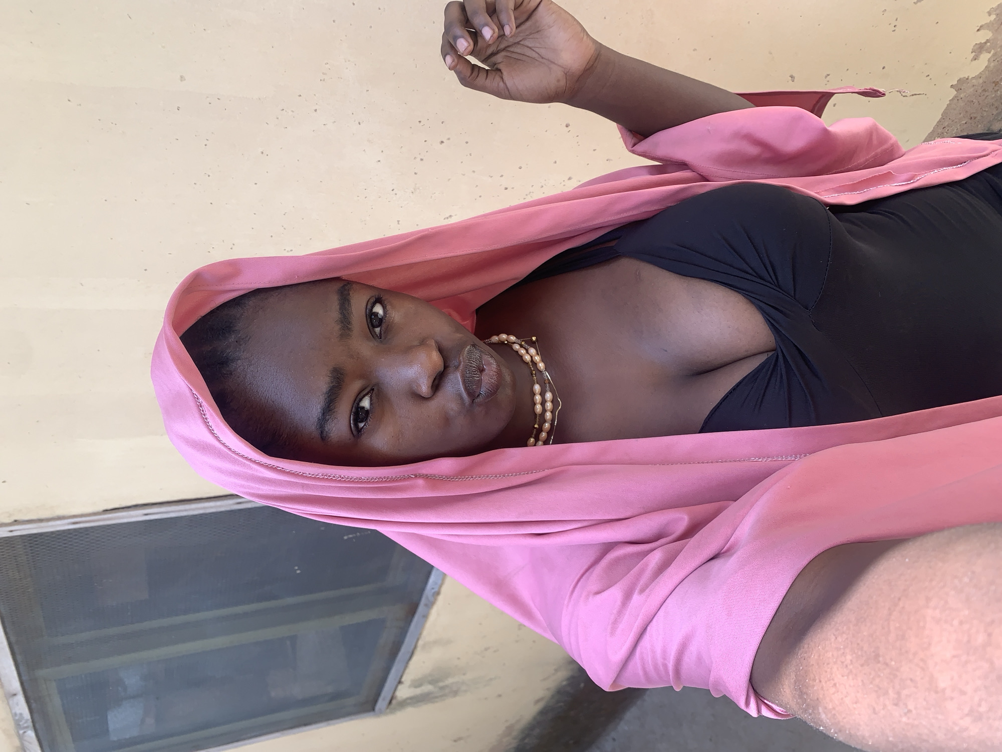 A young woman with dark skin is lying down, wearing a black top and a pink headscarf. She is also wearing a pearl necklace. She is looking at the camera with a slight pout. Her right hand is raised, and she appears to be holding something small between her fingers. She seems to be lying near a window in Rafin Par, Nigeria. The walls are painted a yellowish color.