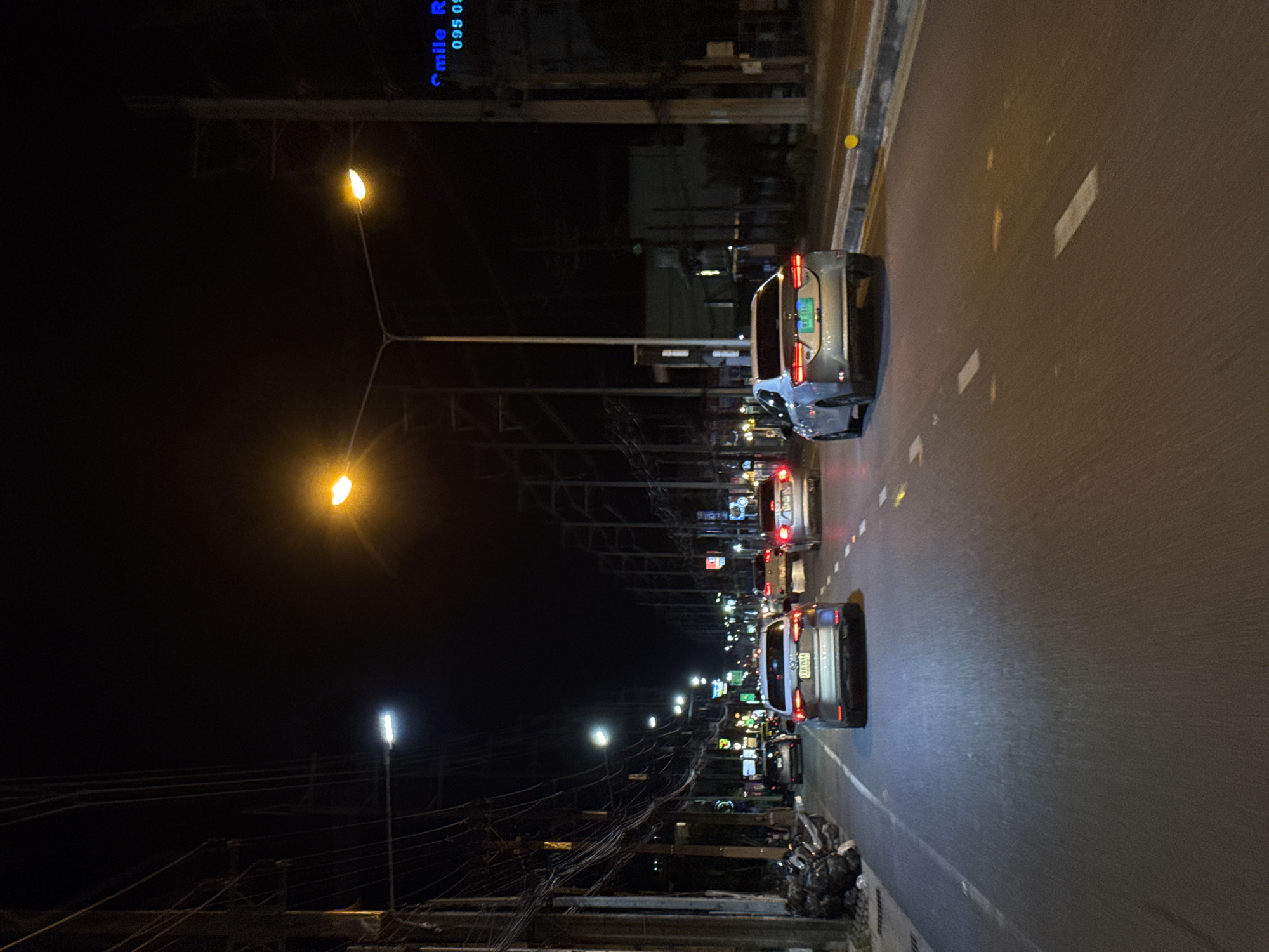 The image depicts a street scene at night in an urban environment. Several cars are driving on a multi-lane road. The street is illuminated by streetlights and the headlights of the vehicles. On the left side of the road, there are power lines and poles, as well as some trash bags piled up. On the right side of the road, there are buildings and signs, one of which has blue lettering that reads "Smile R 095 00". The road surface is asphalt and has visible lane markings. The overall impression is of a typical city street at night with ongoing traffic.