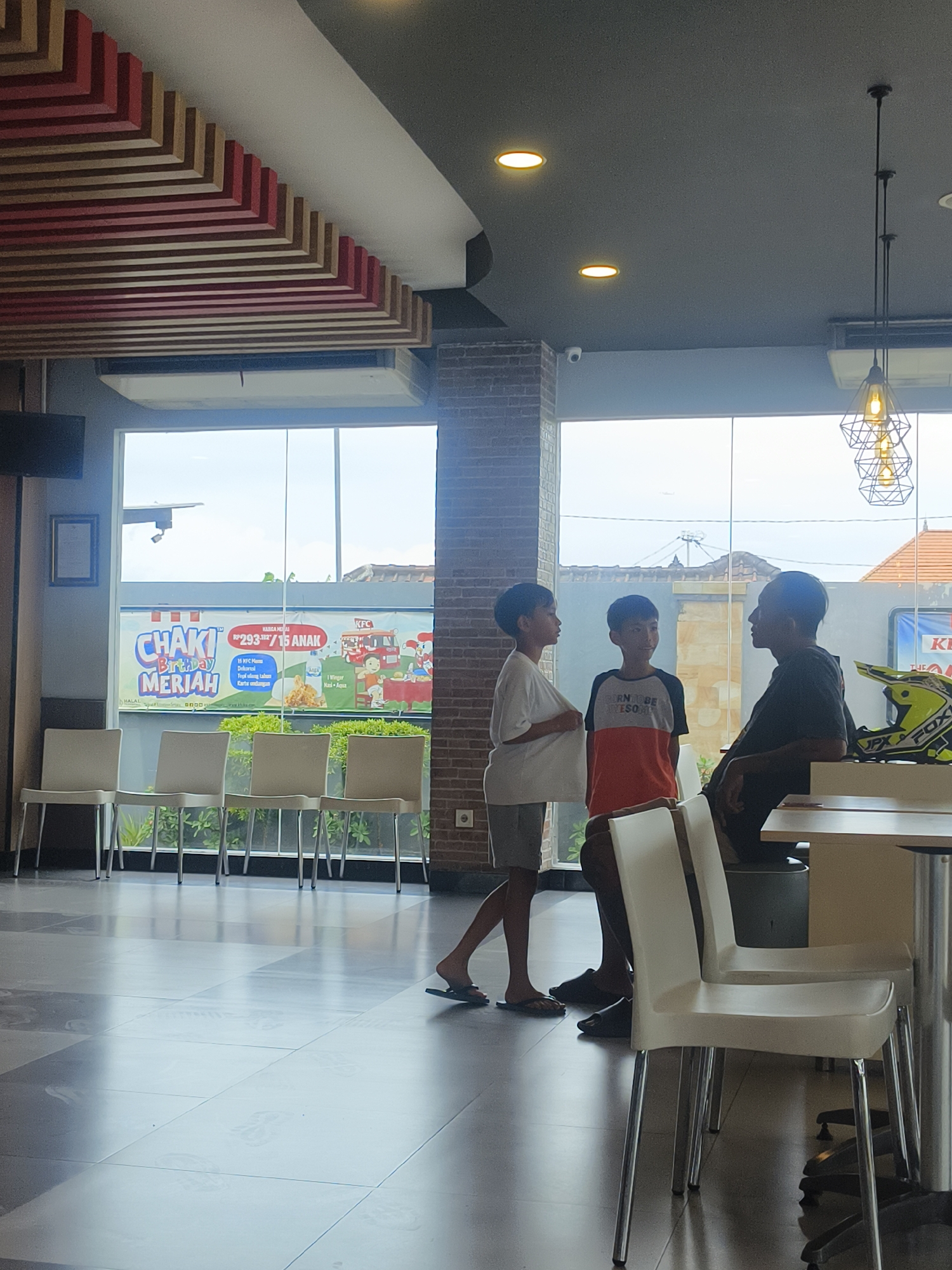 In Denpasar, Indonesia, inside what appears to be a KFC restaurant, a group of three people are standing and conversing near a large window. Two young boys, one wearing a white t-shirt and grey shorts, and the other a t-shirt with red and orange panels, are talking to an adult male dressed in a black t-shirt. The adult is seated or leaning, facing the boys. A yellow and black motocross helmet is visible on a table to the right of the frame. The interior features white plastic chairs lined up against the window, a tiled floor with reflective surfaces, and decorative wooden slats on the ceiling. Through the window, a bright sky and some outdoor structures are visible, including a promotional banner for KFC with "CHAKI Birthday Meriah" and pricing information in Indonesian. The lighting suggests it is daytime.
