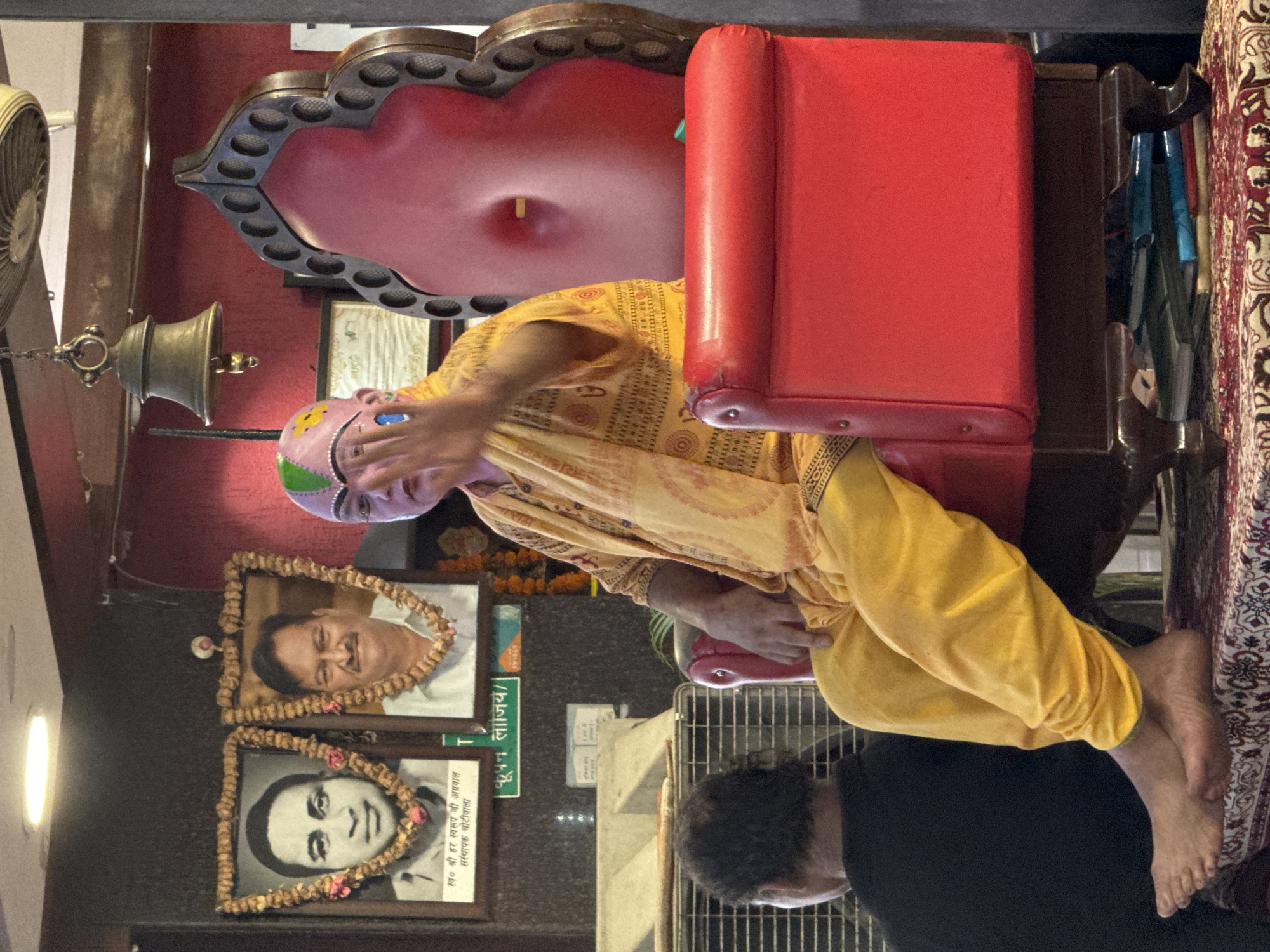 The image captures an indoor scene, possibly a temple or cultural center in Rishikesh, India. In the foreground, a person is seated on a large, ornate red chair. They are wearing a yellow robe and a pink headpiece with green accents. The person's right hand is raised, palm facing forward, in a gesture that could be a blessing or greeting. In the background, two framed portraits hang on a dark wall. The left portrait depicts a man in a white shirt and the right portrait shows a man with a mustache. Between the portraits, a green sign with white lettering is visible. A bell is suspended from the ceiling above and to the right of the portraits. A ceiling fan is also visible in the upper right corner of the image. In the lower left, part of another person's back, wearing a black shirt, is visible. The setting appears to be dimly lit, with a light source illuminating the upper portion of the scene.