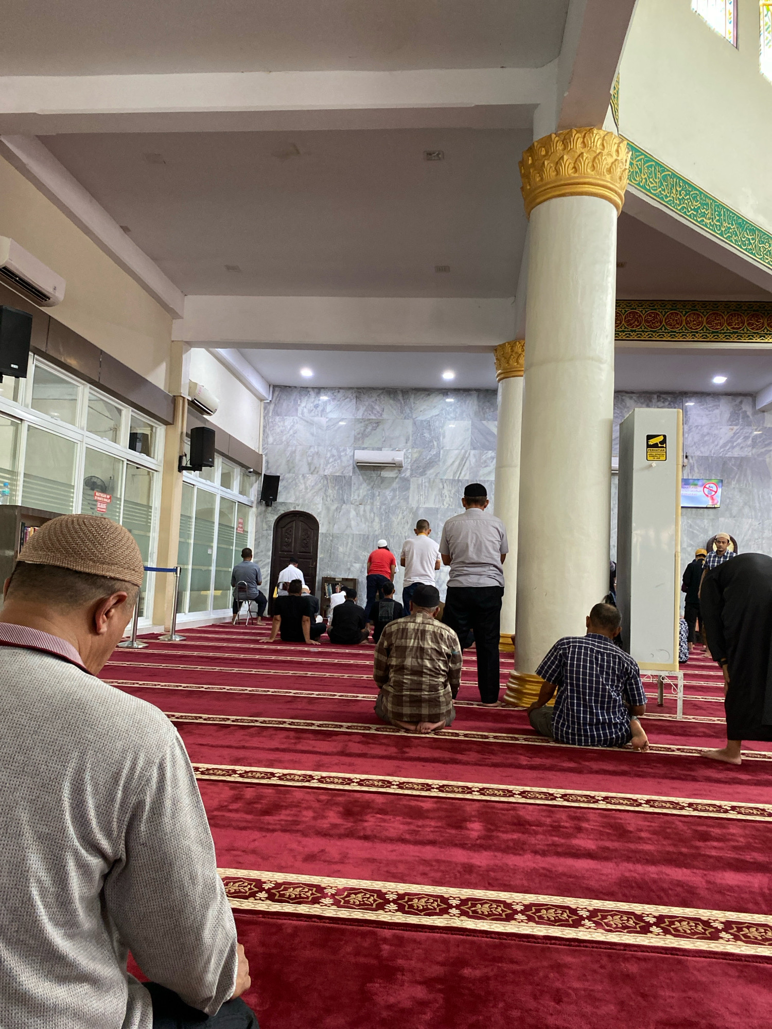 The image depicts the interior of a mosque in Lhokseumawe, Indonesia, likely during prayer time. Several individuals are present, facing the front of the mosque, some kneeling and others seated on a red carpet with intricate golden patterns. The mosque features tall, fluted columns with ornate golden capitals, and the walls appear to be made of marble. A metal detector archway is visible on the right side of the image. The lighting is artificial, and the atmosphere suggests a place of worship.