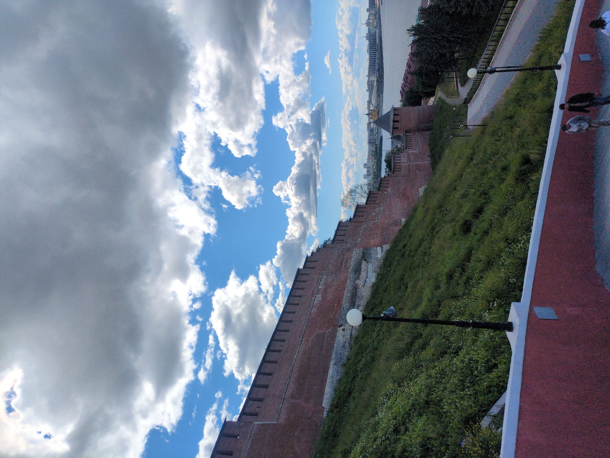 The image captures a scenic view of Nizhny Novgorod, Russia, likely during daylight hours with a dynamic sky featuring both bright blue patches and dramatic, white and grey clouds. The perspective is from an elevated position, looking down and across a landscape.
Dominating the foreground and extending into the midground is a historic brick wall, part of a fortification, which runs diagonally across the frame. The wall is topped with crenellations and appears to be well-maintained, though some sections show signs of age. Adjacent to the wall, a lush green slope is covered in grass and wildflowers, creating a natural contrast to the man-made structure.
A paved pathway runs alongside the wall, and several ornate lampposts are visible, suggesting this is a public area or a promenade. In the distance, a wide river can be seen, with buildings and structures lining its banks, hinting at the city's urban landscape. The architecture of the city in the distance includes what appears to be a Ferris wheel, indicating a recreational area.
A few people are present in the image. On the far right, two individuals are walking along a red-topped path, with white edging, which is also elevated. Further down the path, near the lampposts, another person is visible, seemingly standing or walking. The overall mood is one of peaceful exploration and appreciation of historical and natural beauty. The weather appears to be fair, with the clouds suggesting a possibility of passing showers, but the sunlight breaking through indicates it is not actively raining. The location cues, such as the river and the style of the fortification, strongly point to Nizhny Novgorod. There is no visible text in the image.