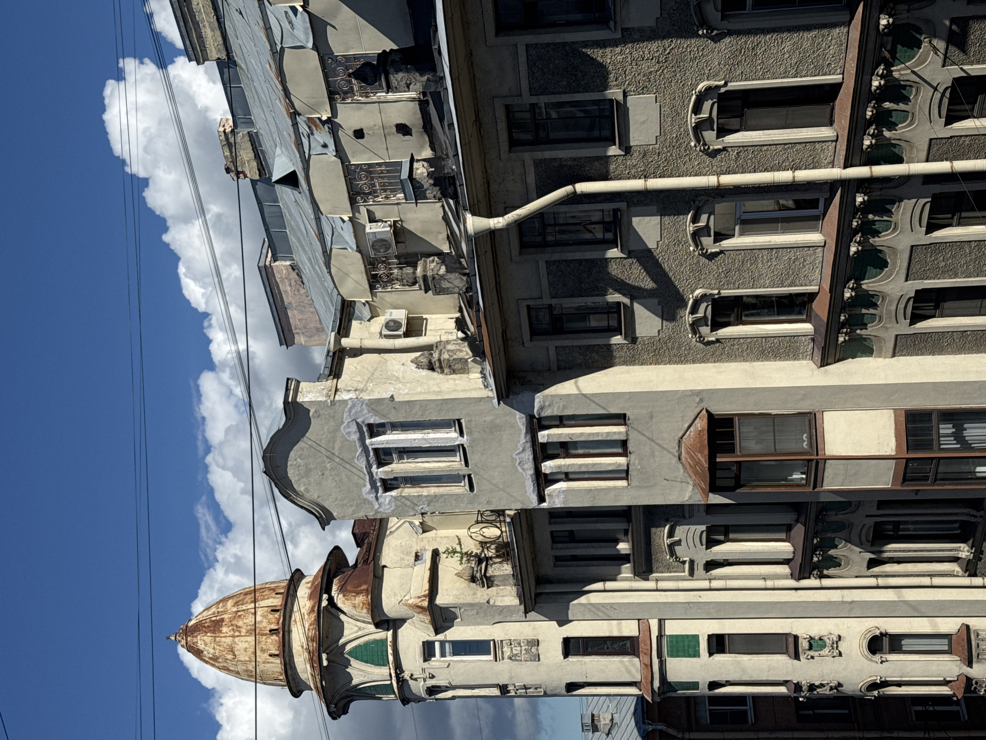 This is a vertical, eye-level shot of an old, ornate building against a bright blue sky filled with dramatic white clouds. The building's facade is a mix of textures and colors, with grey stucco, aged metal roofs, and visible weathering. Numerous windows of various shapes and sizes punctuate the structure, some with decorative detailing.
A prominent feature is a rusted, domed structure in the lower left portion of the image, adding a touch of historical grandeur. Electrical wires stretch across the scene, hinting at an urban setting. The lighting suggests it is daytime, likely late morning or early afternoon, given the strong shadows cast by the building. The weather appears fair and sunny, with the clouds adding visual interest. The architecture suggests a European location, possibly Eastern Europe, with its distinctive style. There are no people visible, and no text is discernible in the image. The overall impression is one of aged beauty and architectural character.