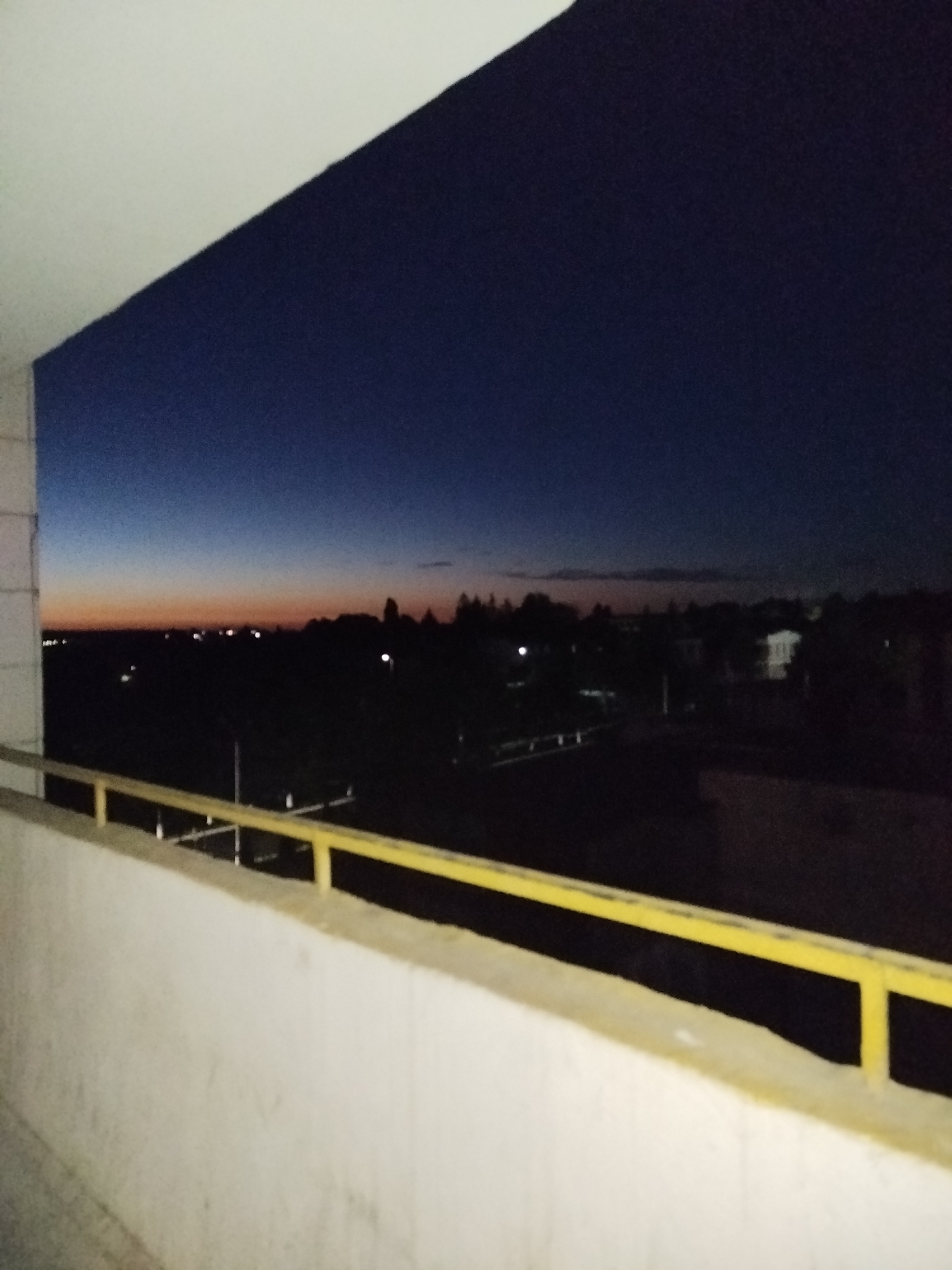 The image shows a view from a balcony at dusk. The sky transitions from a deep navy blue at the top to lighter shades of orange and pink near the horizon, indicating the last light of sunset. Below the sky, a dark silhouette of trees and buildings is visible. Some scattered lights suggest a town or city. In the foreground, a white concrete railing with a yellow metal bar runs horizontally across the bottom of the frame, partially obscuring the view of the landscape. The overall scene is dimly lit and somewhat blurry.