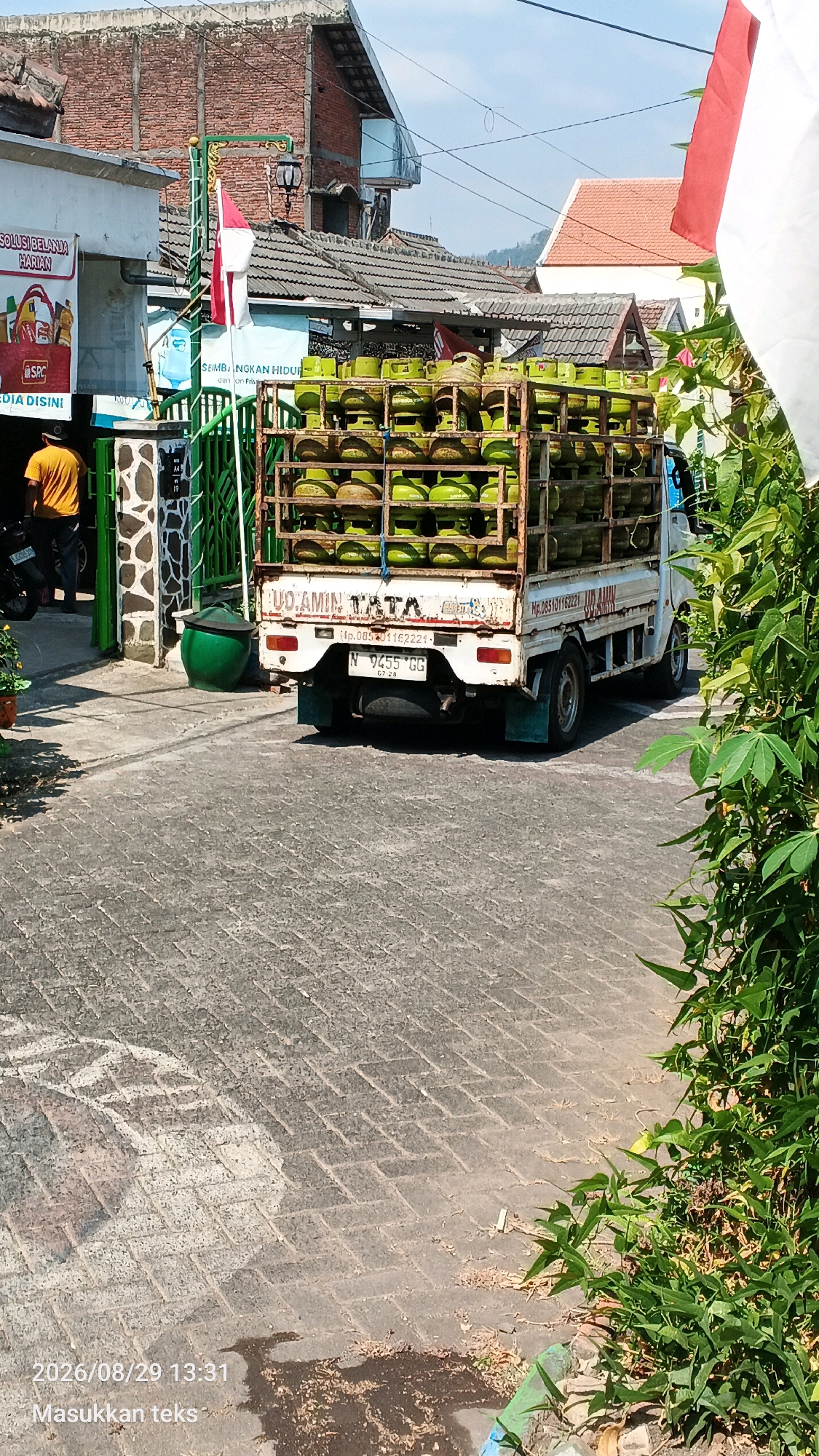 In Sumberporong, Indonesia, a white truck is parked on a paved road, its bed laden with numerous green propane tanks. The truck displays the text "UD AMIN TATA" and a license plate reading "N 9455 CG". To the left of the truck, a building with a white facade has a sign with red and white lettering, and a person in a yellow shirt is visible standing nearby. A green gate with decorative elements stands in front of the building. Red and white Indonesian flags are displayed on a pole near the gate, and a white flag is visible on the right side of the image. Lush green foliage borders the scene on the right. The sky is clear and blue, suggesting a sunny day.