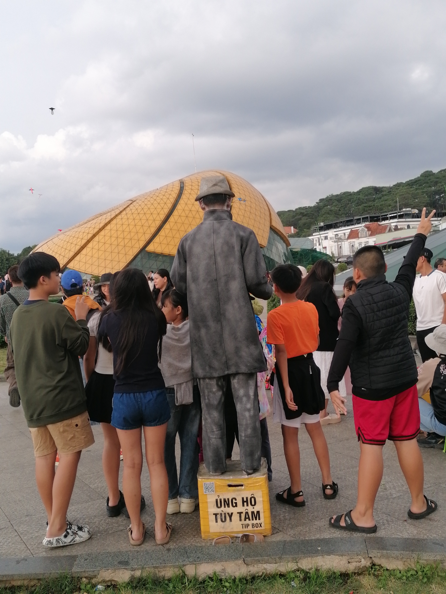 In Da Lat, Vietnam, a group of people gathers around a life-size statue of a man in a suit and hat. The statue is standing on a yellow box labeled "ỦNG HỘ TÙY TÂM TIP BOX" (Donation as you wish, Tip Box).
The statue is positioned facing away from the camera, its back to the viewer, with its right arm raised in a peace sign gesture. A boy in an orange t-shirt and black shorts stands to the left of the statue, while another boy in a green long-sleeved shirt and khaki shorts stands to the far left.
Behind the statue, a woman in a black t-shirt and blue denim shorts is visible, along with other individuals. In the background, a large, golden, dome-shaped building with a distinctive architectural design dominates the scene. The sky above is overcast.
Further in the distance, a lush green hillside is dotted with buildings, suggesting an urban or semi-urban environment. Two kites are visible in the sky, adding a touch of playful activity to the scene. The ground is paved with concrete, and a strip of grass is present in the foreground. The overall atmosphere appears casual and public, possibly a tourist attraction or a public gathering space.