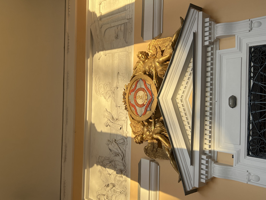 This image depicts a detailed section of the Mikhailovsky Palace (State Russian Museum) facade in Saint Petersburg, Russia. The scene focuses on an ornate architectural element above an entrance, illuminated by direct sunlight from the upper right, casting sharp shadows.
At the center, two golden, winged female figures, styled as Victories or angels, recline symmetrically atop a classical pediment. They support a prominent, circular gilded shield featuring a central red disc with a crowned monogram "E.S." (for Grand Duchess Elena Pavlovna), framed by concentric silver and gold rings with decorative patterns. The pediment itself is white, with classical cornices and dentil moldings.
Immediately below the pediment, a white architectural frame for a door or large window is visible. It includes fluted pilasters on the sides and a decorative architrave. An oval-shaped, translucent light fixture is mounted above a dark, intricate metal grille that partially covers the opening.
Above the golden figures and shield, a long, rectangular bas-relief panel is integrated into the wall. This relief, rendered in a light grey or white material, depicts multiple human figures in various poses, some standing and some kneeling, engaged in a narrative or allegorical scene. To the right of this primary bas-relief, another architectural element, possibly a classical archway or building fragment, is sculpted in relief.
The building wall is painted a uniform, light ochre or pale yellow. The upper portion of the image shows a plain, light-colored ceiling or overhang, partially shadowed. The strong sunlight highlights textures and details on the right side of the elements while creating deep, angular shadows across the left. No living individuals are present; all depicted figures are sculptural.