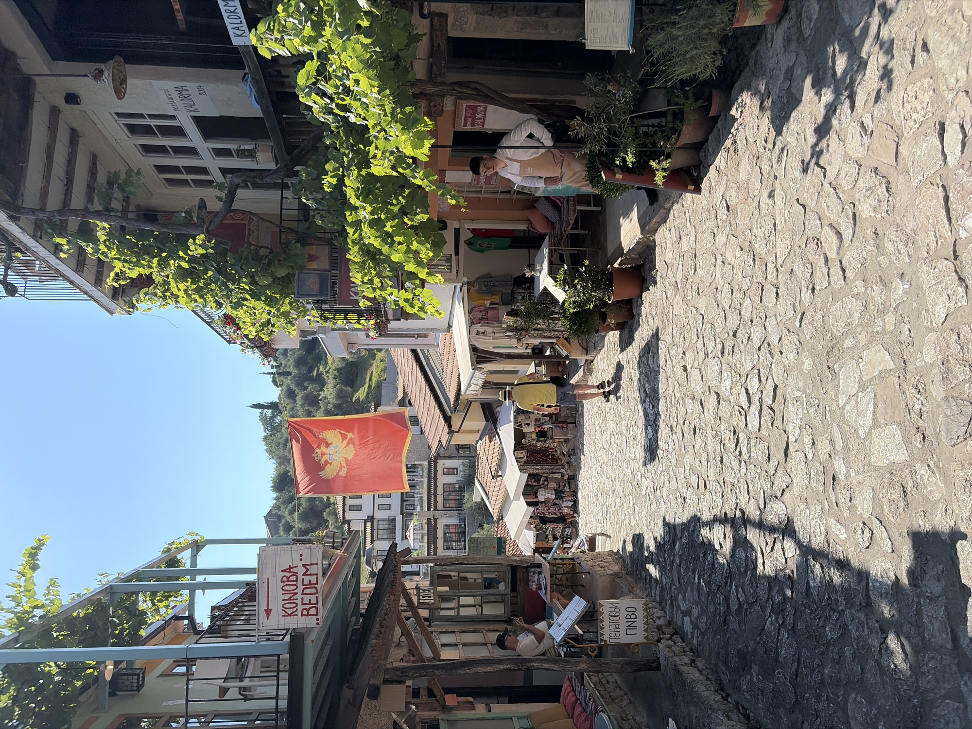 The image depicts a sun-drenched, cobblestone street in Stari Bar, Montenegro. The street slopes uphill, lined with old stone buildings that house shops and restaurants. Green vines drape over balconies and trellises, adding to the Mediterranean charm.
On the left side of the street, a sign with an arrow points to "KONOBA BEDEM." Below this, another sign reads "ГРАНАТОВЫЙ ПИВО" (Pomegranate Beer in Russian). A person is seated at a small table outside this establishment, looking at their phone. Cushions are arranged on a bench, and there are potted plants.
Across the street, a vibrant red flag with a gold crest, the flag of Montenegro, hangs from a building. Further up the street, another sign indicates "restaurant KALDRMA." People are visible browsing stalls selling souvenirs and clothing, with white umbrellas providing shade. One individual in a yellow shirt and blue shorts walks away from the camera, and another person wearing an apron stands outside a shop. The architecture features stone facades, wooden beams, and tiled roofs. The sky is clear and blue.