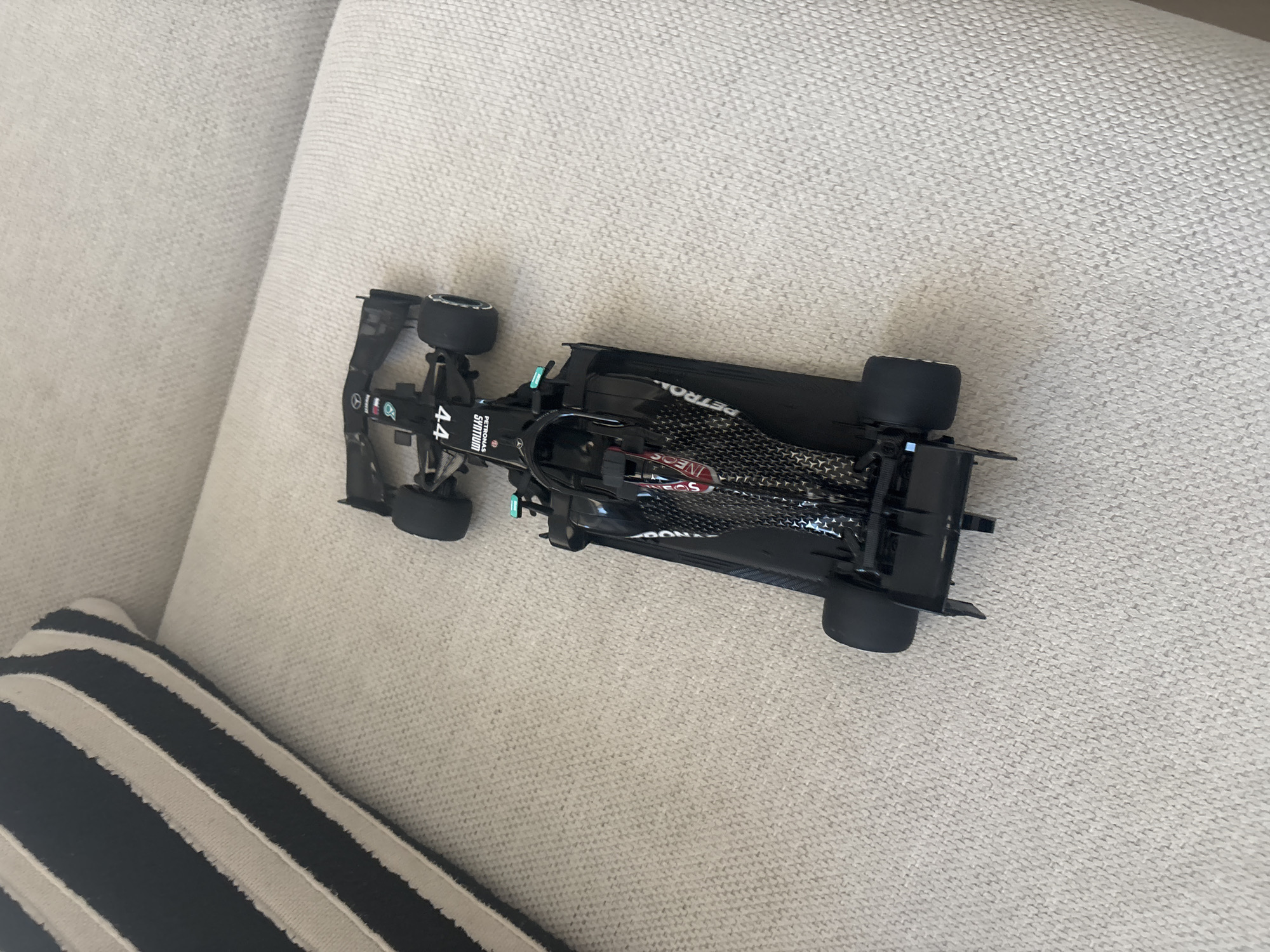This is a top-down, angled shot of a black Formula 1 toy car, likely a model of a Mercedes-AMG Petronas F1 W11 EQ Performance car, placed on a textured, light-colored fabric surface, possibly a sofa or carpet. The car is positioned diagonally in the frame, with its front pointing towards the top left.
The car is detailed, featuring a sleek black body with prominent sponsors' logos visible. Text on the car includes "PETRONAS", "SYNTIUM", "44" (likely referencing Lewis Hamilton's car number), and "INEOS". The car has visible aerodynamic elements, a detailed cockpit, and black tires.
In the bottom left of the frame, a portion of a striped cushion or pillow is visible. The cushion has alternating wide stripes of dark blue/black and off-white. The fabric of the main surface is a woven textile with a subtle pattern, and there are a few small dark specks visible on it, suggesting it might be a carpet or upholstered furniture.
The lighting appears to be natural daylight, casting soft shadows, and the overall scene suggests an indoor setting. There are no people visible in the image. The mood is static and observational, focusing solely on the toy car and its immediate surroundings. The location context of Dubai, United Arab Emirates, does not offer any specific visual cues within the image itself, as the scene is generic and could be found in many indoor environments.