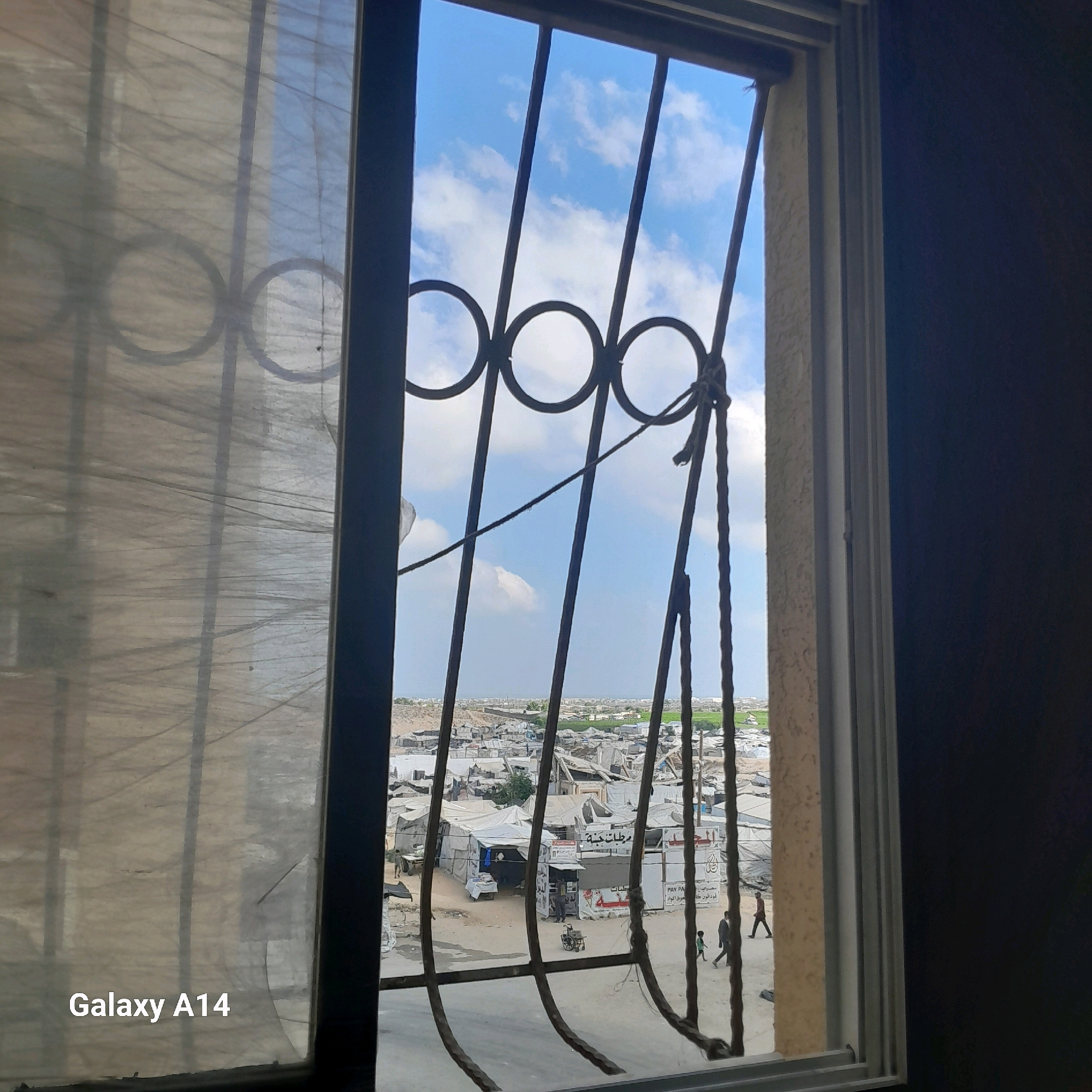 This image, taken in the city of Khan Yunis, Palestinian Territories, presents a view through a barred window overlooking a densely populated area. The foreground features a sheer, translucent curtain partially obscuring the view. Beyond the curtain, a black metal grate with circular accents is visible, with a rope tied to it. The main scene depicts a landscape of makeshift structures, predominantly tents and simple buildings, suggesting a refugee camp or a densely populated informal settlement. The sky above is a clear blue with scattered white clouds. In the middle ground, several small stalls or shops with signage are visible, and a few individuals are present. One person is near a wheelchair, and others are walking. The overall impression is one of a busy, lived-in community. The text "Galaxy A14" is watermarked in the bottom left corner.