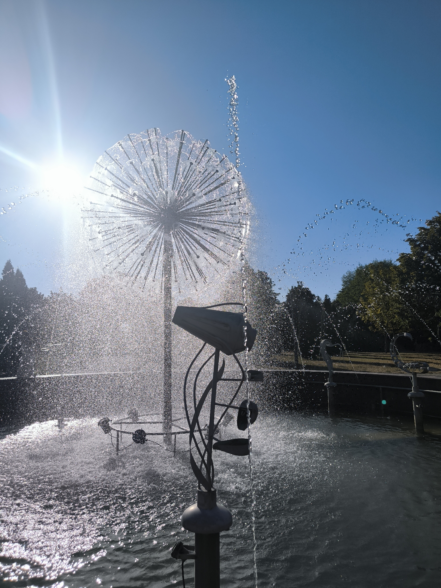 The image shows a close-up of a modern dandelion-shaped fountain in a park, likely in Chișinău, Moldova. The sun is shining brightly from the upper left, creating a lens flare and illuminating the water droplets spraying from the fountain. The fountain itself is a metallic sphere with many thin rods radiating outwards, from which water is being sprayed in a delicate, mist-like fashion. In the foreground, the base of the fountain is visible, with some curved metal elements. The water is spraying upwards and outwards, creating arcs and a fine mist that catches the sunlight. In the background, there are trees, indicating a park-like setting. The sky is clear and blue. The overall impression is one of a bright, sunny day with the fountain being the central focus. The water appears to be falling into a pool or basin.