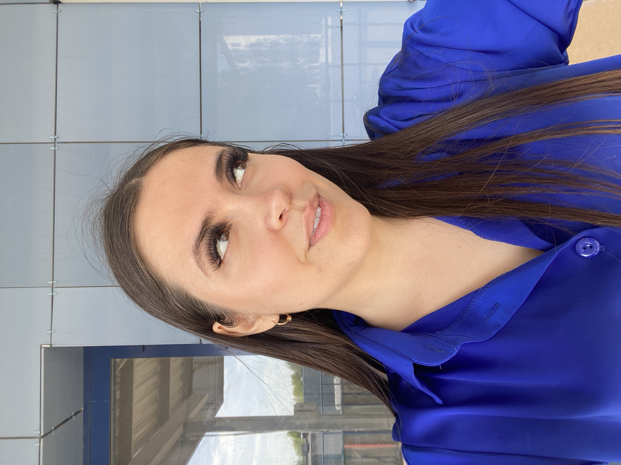 The image shows a young woman with brown hair, wearing a bright blue button-up shirt. She is positioned in front of a wall composed of square, light gray panels, possibly made of glass or a similar material. The backdrop includes what seems to be a window or a glazed section, with a view revealing elements like sky and vegetation, suggesting an outdoor location. The image appears to be taken in Chaikovsky, Russia, as inferred from external context.