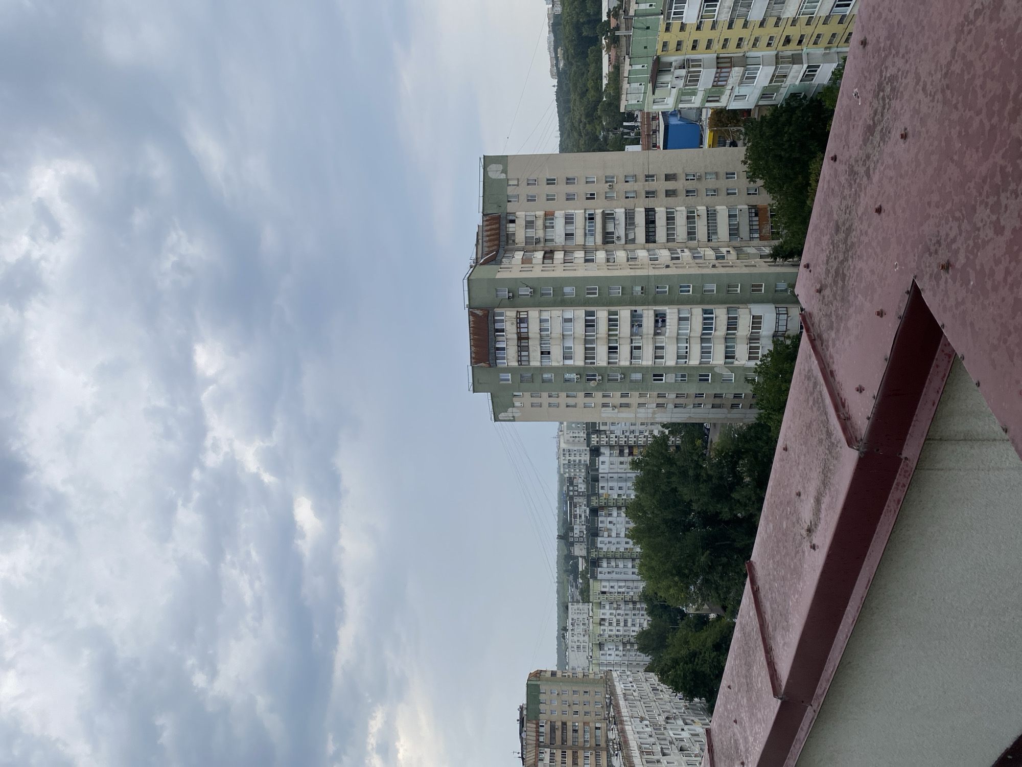 This is an exterior, high-angle, medium shot taken from a rooftop overlooking a dense urban neighborhood. The sky is overcast with a heavy layer of gray clouds. In the foreground, the edge of a reddish-brown metal roof with visible bolts and scuff marks is prominent. Beyond this, a panorama of multi-story apartment buildings fills the midground and background. These buildings are primarily light-colored, with accents of green and yellow on some facades. Many feature rows of balconies and air conditioning units. Trees, mostly green and leafy, are interspersed between the buildings, suggesting a temperate climate. In the distance, a faint horizon line with some structures and trees is visible. The scene appears to be in Chișinău, Moldova, a city known for its uniform apartment block architecture. There are no people or vehicles visible in the frame.