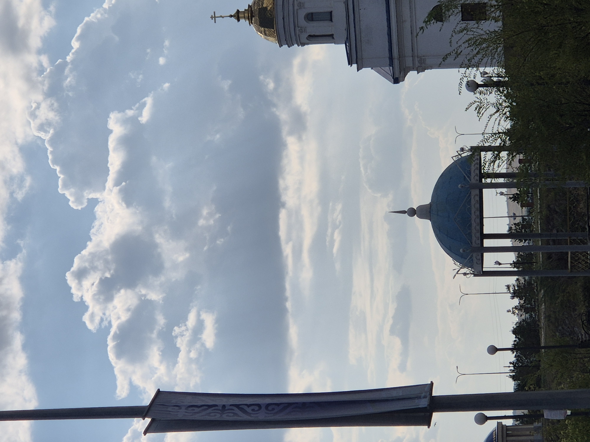 The image captures a view of Kokshetau, Kazakhstan, with a prominent sky filled with dynamic, white cumulus clouds against a pale blue background. In the foreground, a tall, dark gray pole with a faded banner attached on the left side dominates the left edge of the frame. The banner features a decorative, repeating pattern in a lighter shade. To the right, a white building with a golden dome, topped with a cross, rises into the sky, suggesting it is a church or religious structure. Below the dome, several arched windows are visible on the building's facade. In the middle ground, a blue-domed gazebo with intricate metalwork is situated, and beyond it, a line of dark green trees forms a backdrop. Several streetlights with spherical lamps are also visible scattered throughout the landscape. The overall scene suggests an outdoor public space, possibly a park or a plaza, under a partly cloudy sky.