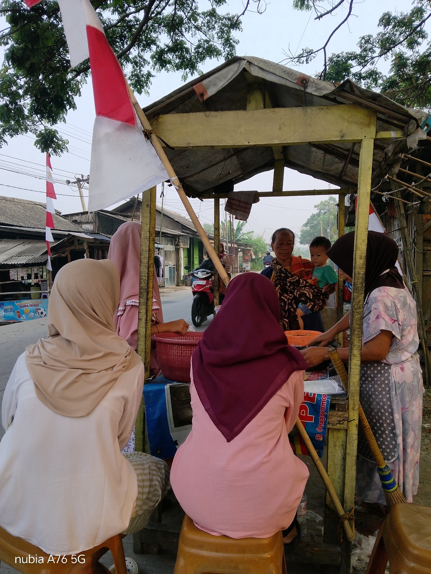 In Cakung, Indonesia, a street vendor operates from a makeshift stall. A red and white Indonesian flag flies prominently above the structure. Three women, all wearing hijabs, are visible at the stall. One woman, in the foreground and from behind, wears a beige hijab and a white shirt. To her right, another woman, also seen from behind, wears a maroon hijab and a pink shirt. The third woman, partially visible on the right side of the frame and facing the stall, wears a dark hijab and a patterned dress with polka dots. She appears to be interacting with an older woman who is holding a baby. The older woman, dressed in a patterned cloth and an orange sash, stands behind the stall. The stall itself is constructed with rough wood and covered with a tarp. Various items are displayed for sale, including what appears to be packaged goods and a red basket. A motorcycle is parked on the street behind the stall. The scene suggests a bustling local marketplace.