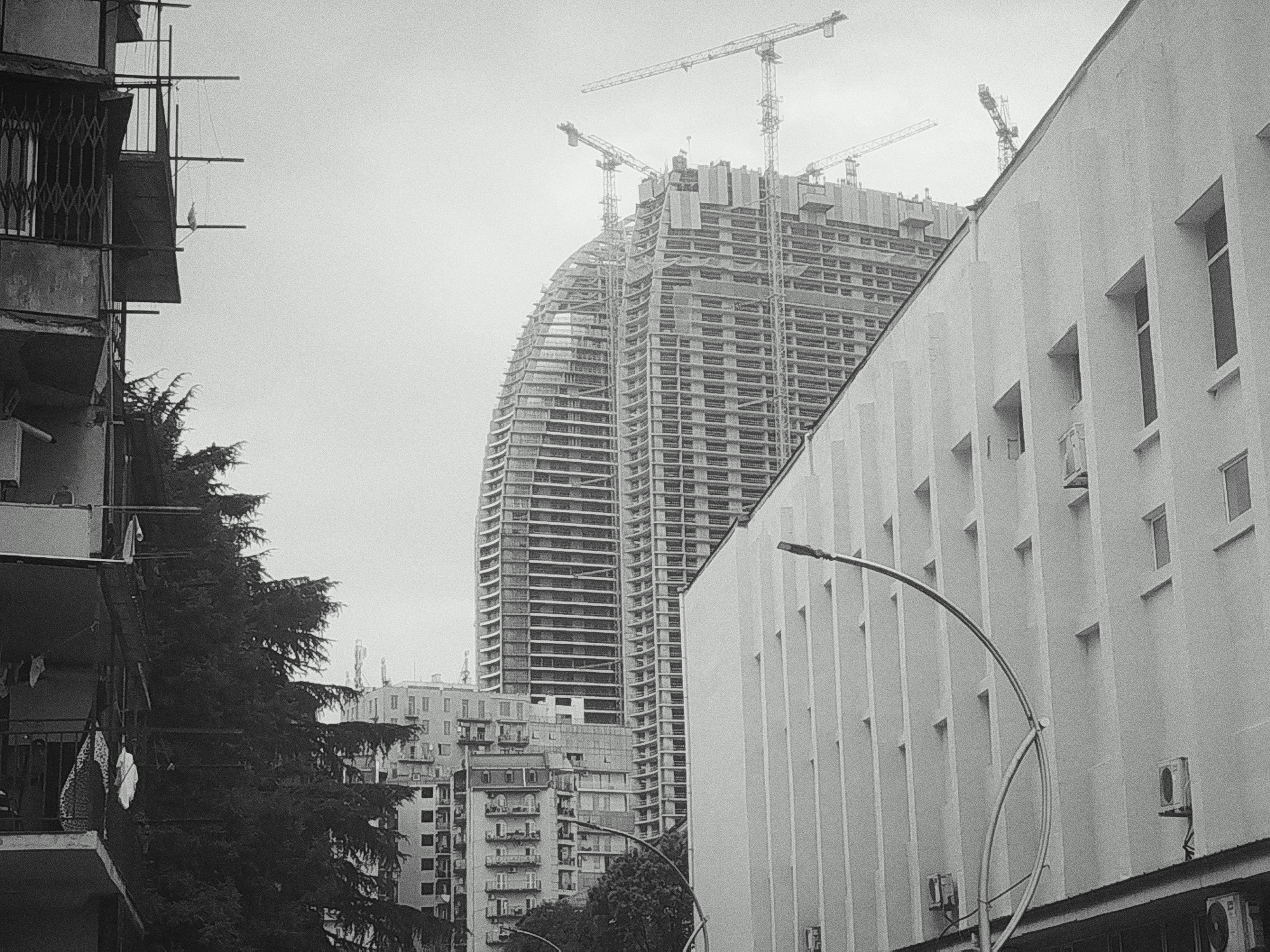 The black and white image depicts an urban scene in Batumi, Georgia, dominated by a towering skyscraper under construction. The unfinished structure, with its exposed rebar and concrete framework, reaches towards a cloudy sky, and several construction cranes are visible atop its highest levels. To the right of the skyscraper, a multi-story building with a light-colored facade and regularly spaced windows runs parallel to the street. On the left side of the frame, an older, multi-story apartment building with balconies and barred windows is partially visible. A large, dark evergreen tree stands between the apartment building and the construction site. Laundry hangs from lines on a balcony of the apartment building. The overall impression is one of urban development, with a stark contrast between older and newer construction.