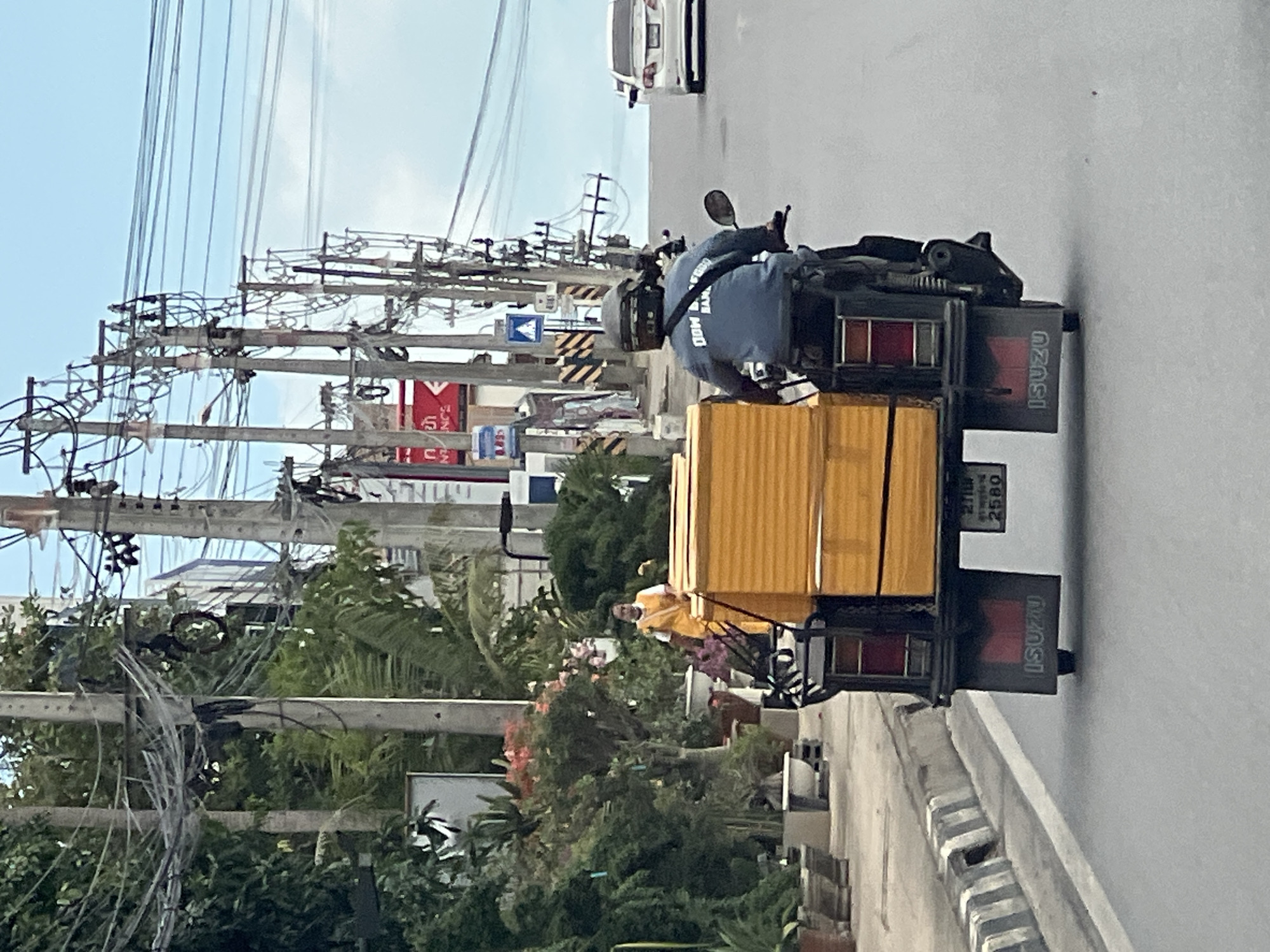 In Ban Bang Rak, Thailand, a motorcyclist wearing a helmet and a blue long-sleeved shirt with text on the back is riding a three-wheeled motorcycle loaded with stacked yellow items. The motorcycle has an "ISUZU" logo on its rear. The scene is on a road with a concrete curb and painted white lines. On the left side of the road, there is a sidewalk with potted plants and a woman in a yellow outfit. Utility poles with numerous wires are visible along the road, and a white car is in the distance. The sky is blue with some clouds.