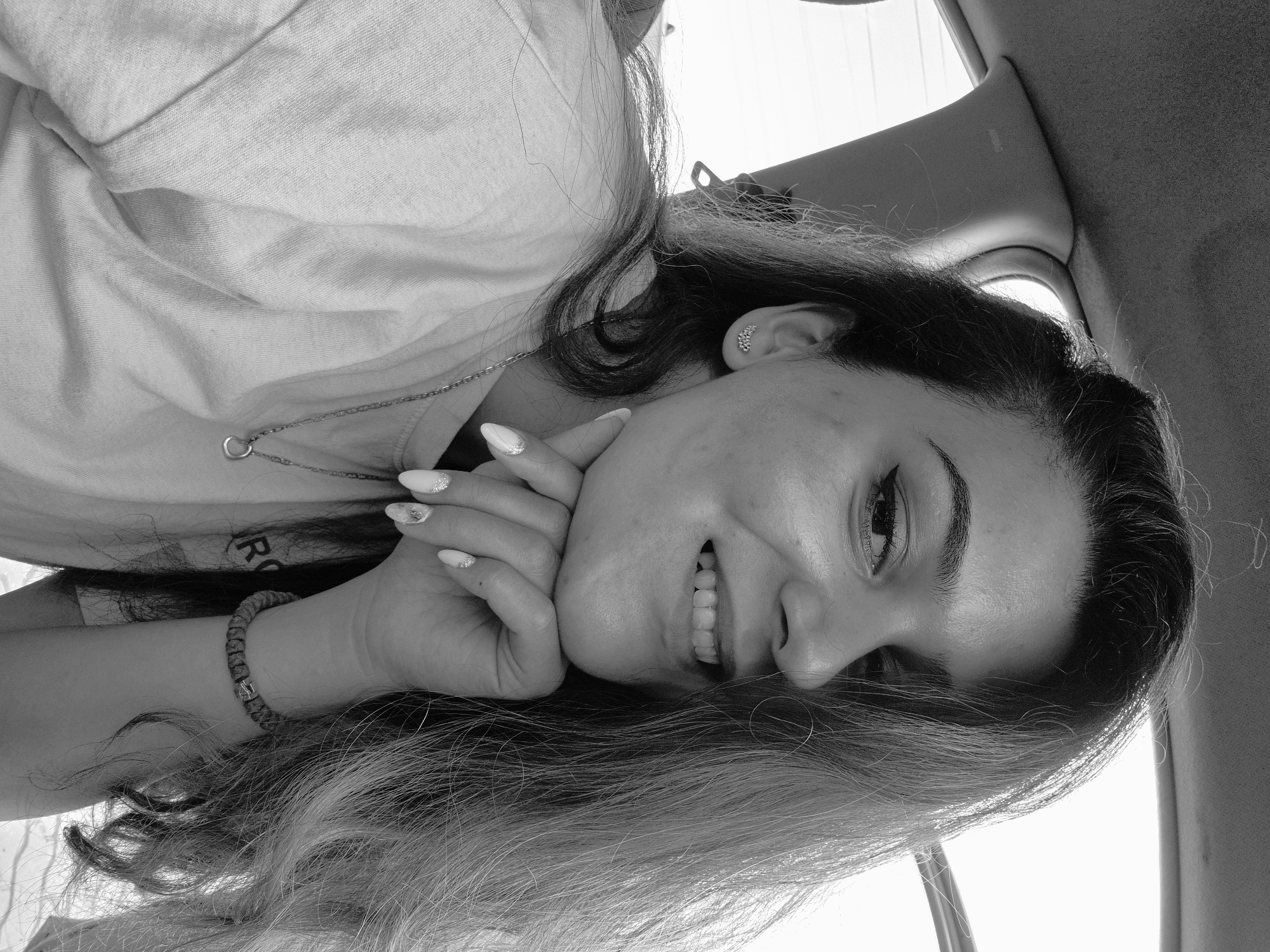 A black and white, low-angle portrait features a young woman smiling brightly, presumably inside a vehicle. She has long, dark hair with lighter undertones or highlights, framing her face. Her hands are gently clasped beneath her chin, showcasing manicured nails with a light polish and glittery accents. She wears a light-colored top, on which a partial letter "R" is visible. Adorning her are a necklace with a small heart-shaped pendant, cluster earrings on her right ear, and a braided bracelet on her right wrist.
The woman's expression is joyful and engaging, characterized by a wide, genuine smile that reveals her teeth. Her eyes appear bright, and her dark eyebrows are well-defined. The setting indicates the interior of a car or similar vehicle, with a faint "AIRBAG" label visible on the ceiling or upper pillar. Bright daylight streams from above and behind her, suggesting a daytime scene. The overall mood of the image is cheerful and relaxed. While the provided context places the location in Sofia, Bulgaria, no specific visual cues within the image confirm this detail.