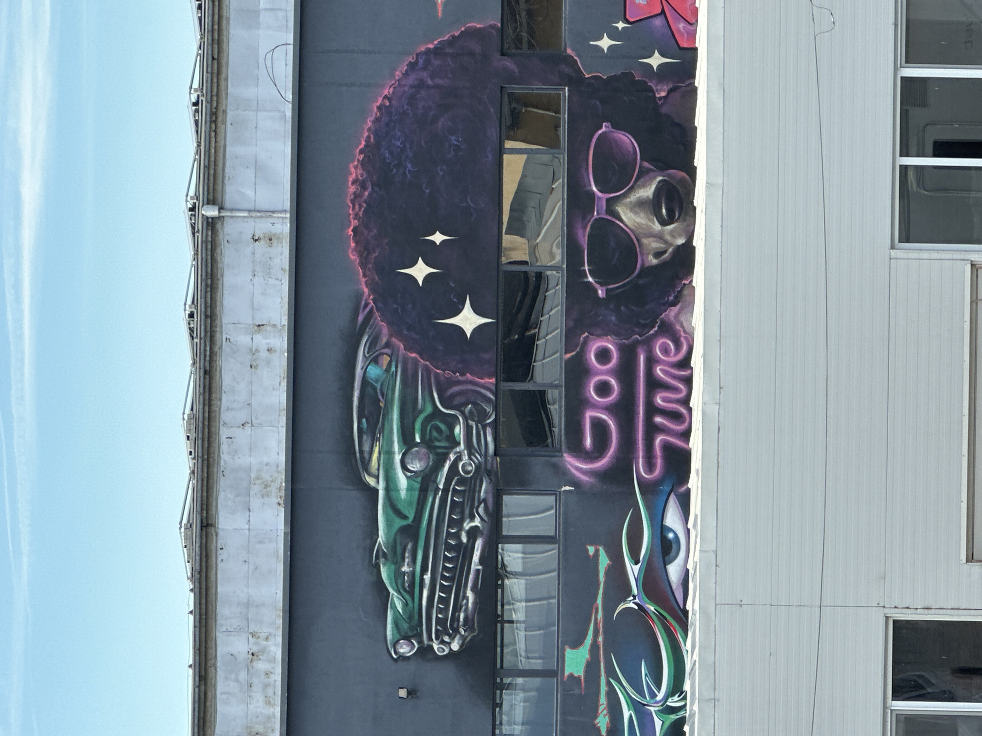 A mural on the side of a building in Sofia, Bulgaria, features a retro-style illustration incorporating elements such as a green classic car, a character with an afro and sunglasses, stars, and neon lettering. The mural wraps around the windows of the building. The building is located next to a light blue sky.