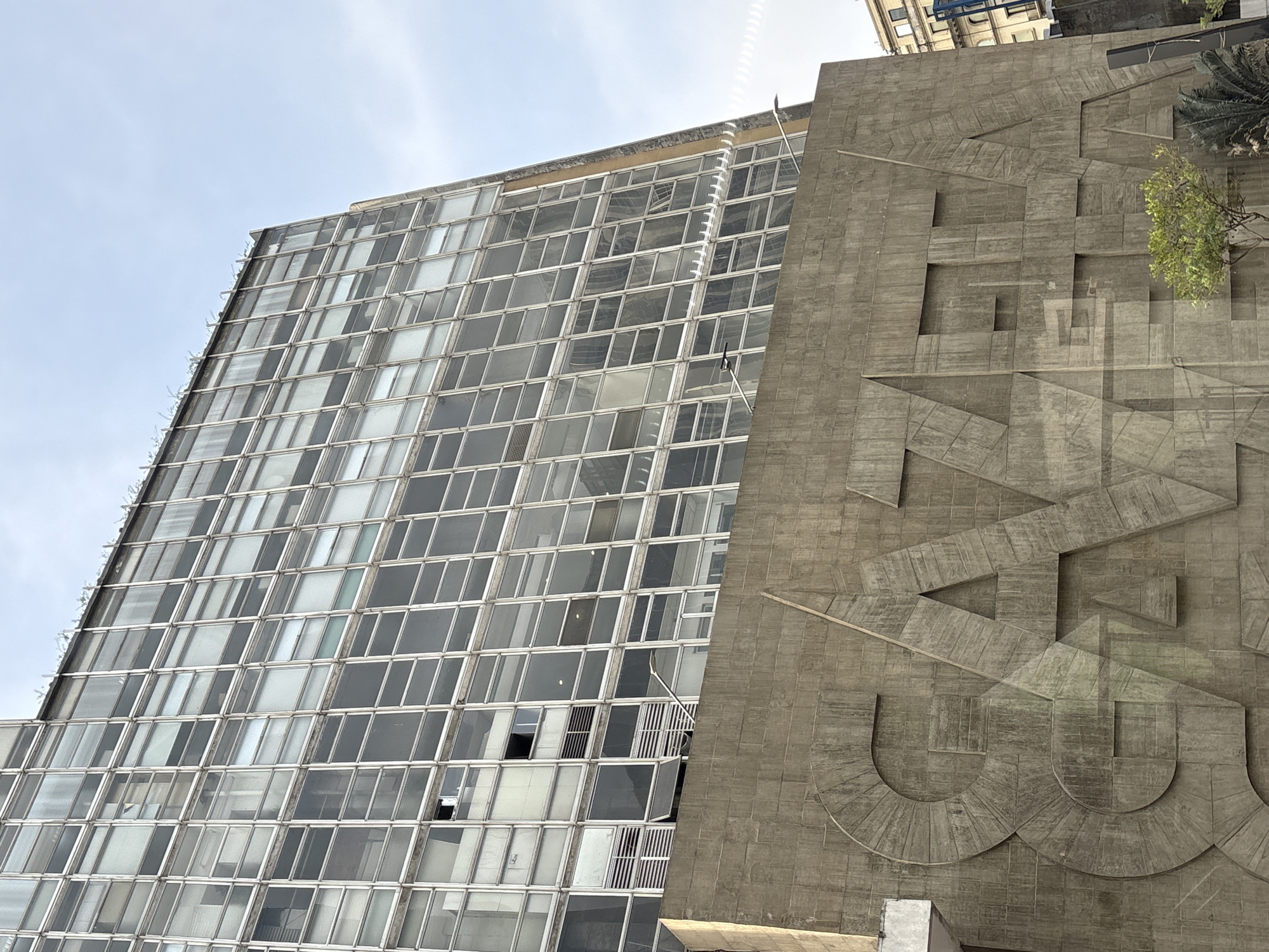 The image shows the building of the newspaper "Gazeta" located in São Paulo, Brazil. The building features a modern architectural style with a facade composed of numerous glass windows. The lower part of the building has a concrete base with large, relief lettering that spells out "GAZETA". The lettering is geometric and three-dimensional, creating a striking visual element. The sky is overcast and the overall lighting suggests it is daytime. There are some trees and a palm tree in front of the concrete base. The image captures a low-angle view of the building, emphasizing its height and architectural design.