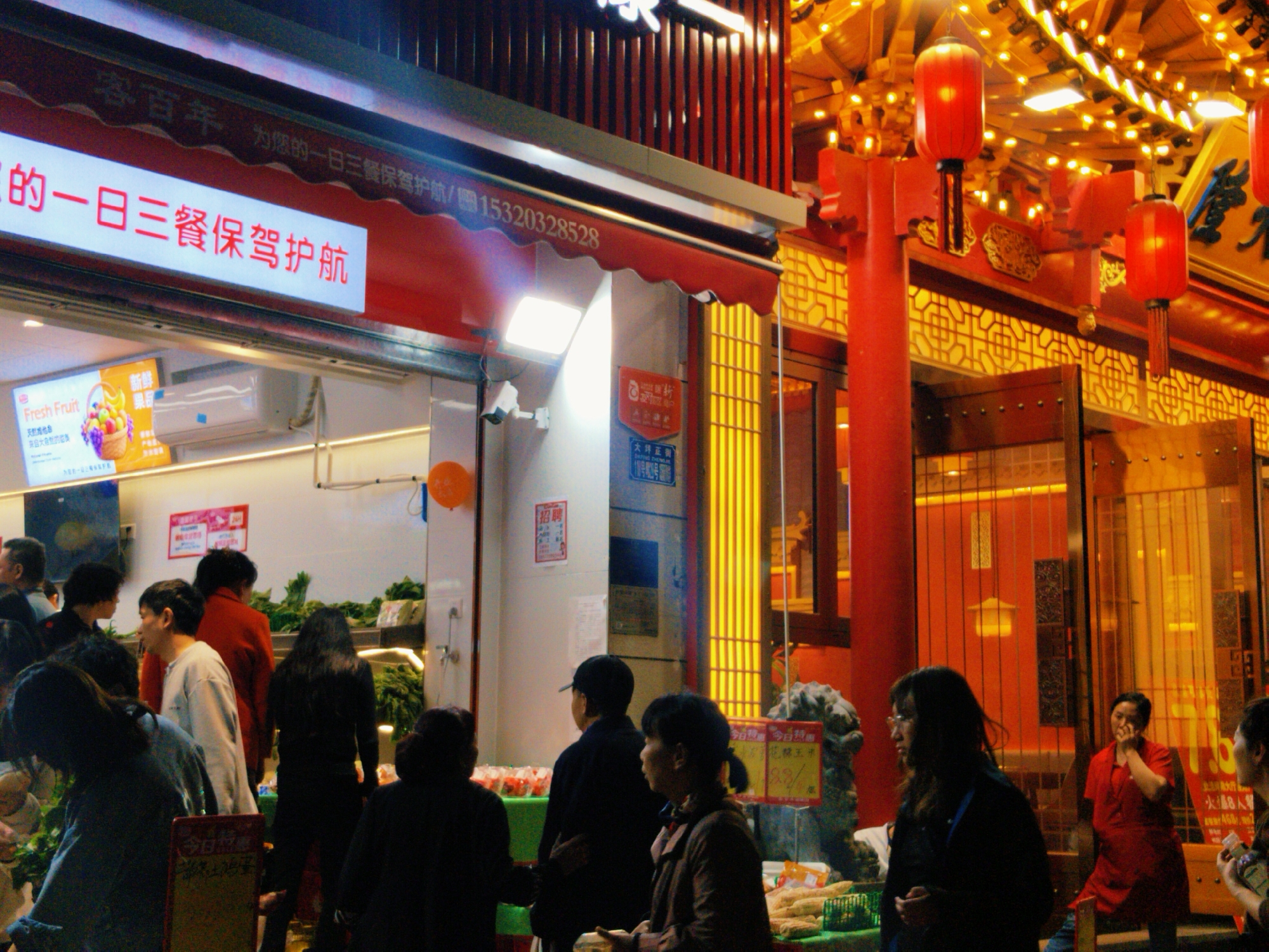The image shows a street scene in the Yuzhong District of Chongqing, China, at night. A bustling shop front featuring fruit and vegetables is visible, with customers milling about, examining and purchasing goods. The shop has a red awning with white text in Chinese. Above the awning, a sign displays Chinese characters. Inside, there is a display of "Fresh Fruit." The interior is illuminated by bright lighting.
Several people are visible. Some are browsing the produce, while others are waiting. An individual stands near the entrance wearing a red shirt. Another woman is seen in a black jacket. The people in the image are dressed in various attire, suggesting a variety of ages and backgrounds.
To the right of the shop front, traditional Chinese architecture is on display, with red columns, patterned window frames, and hanging lanterns. The lanterns add a warm glow to the scene. A stone statue sits to the right of the open entrance with signs.
The scene is busy and vibrant, capturing the essence of an active market in a Chinese city. The photograph is taken at night.