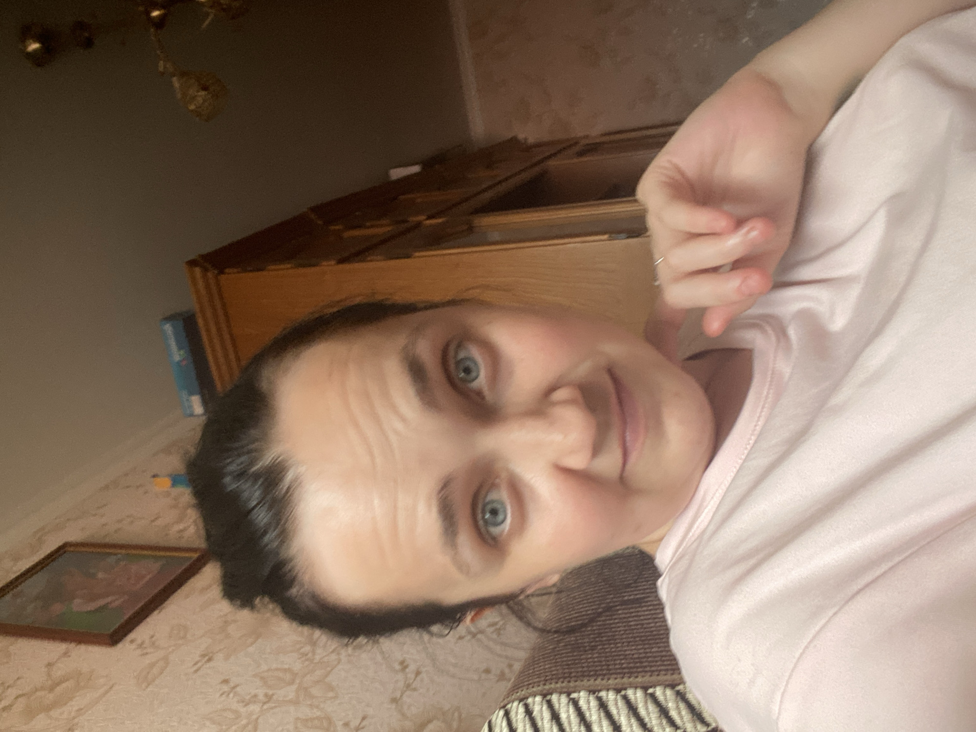 A medium-shot, waist-up, selfie-style photograph shows a Caucasian woman with dark hair pulled back and blue eyes. She is wearing a light pink t-shirt. Her left hand is raised and resting near her face, with a ring on her right index finger. The background appears to be a home interior, with a patterned wallpaper on the left, a wooden cabinet or bookshelf in the center, and a light fixture visible on the upper right. The lighting is dim, suggesting an indoor setting.
