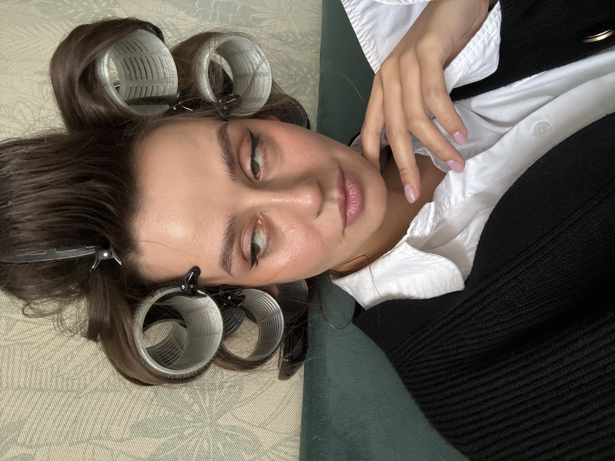 A woman is lying on a surface, possibly a bed or a couch. She has brown hair with several hair rollers and clips in it. She is wearing a white collared shirt with a black sweater on top, and her hand is resting near her neck. The background includes a patterned surface underneath her head and a green surface beneath her body. There is no indication of this image being taken in Krasnoyarsk, Russia.