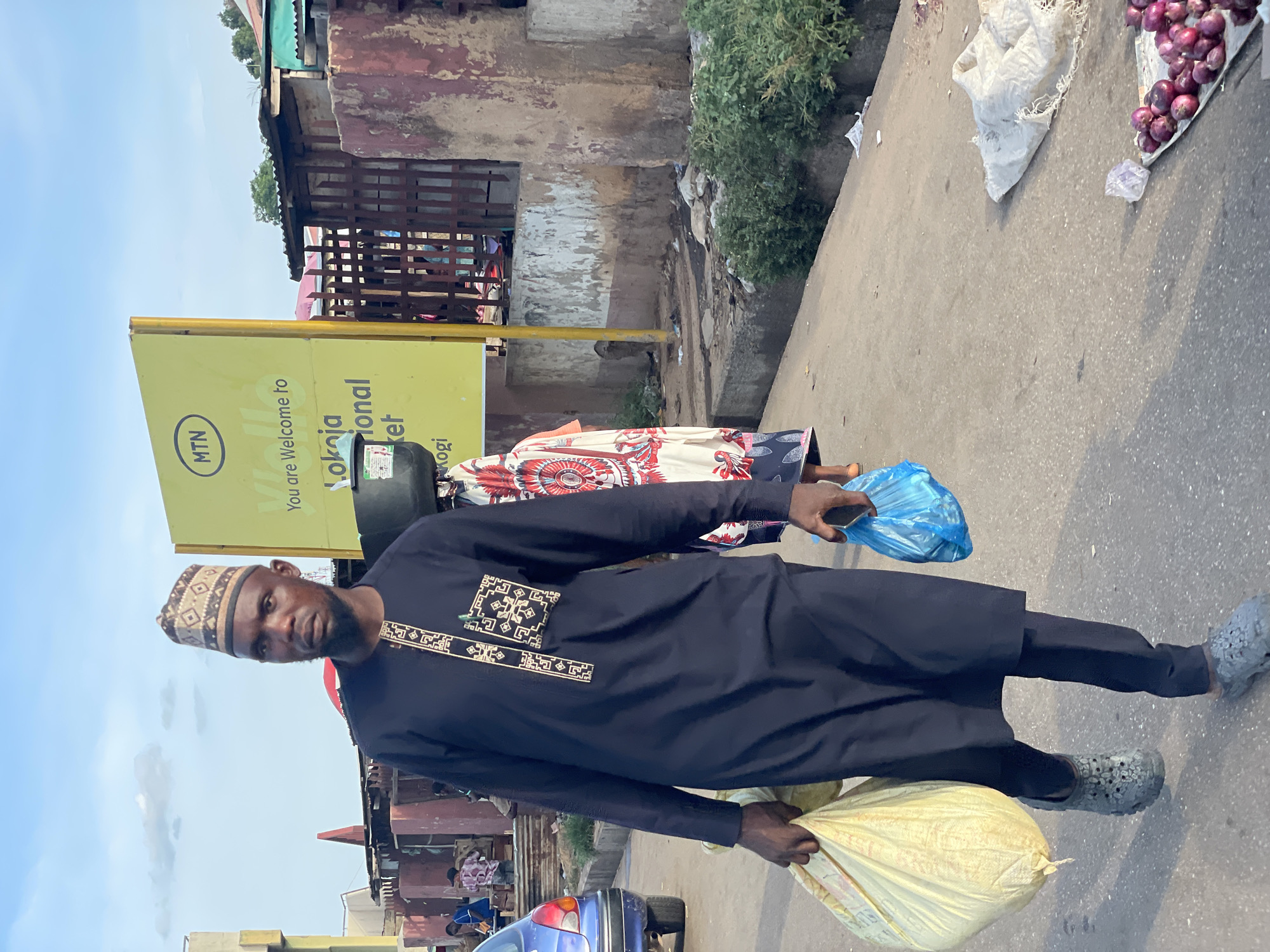 A man in a dark, embroidered tunic and patterned cap is standing on a street in Lokoja, Nigeria. He holds a blue plastic bag in one hand and a yellow cloth bag in the other. He is standing next to a yellow sign that says "You are Welcome to Lokoja International Market," with the MTN logo. Behind him, there is a building with deteriorated paint and exposed wooden structures. In the background, there's a sidewalk with loose debris and a tray of red onions for sale. A blue car is also partially visible.